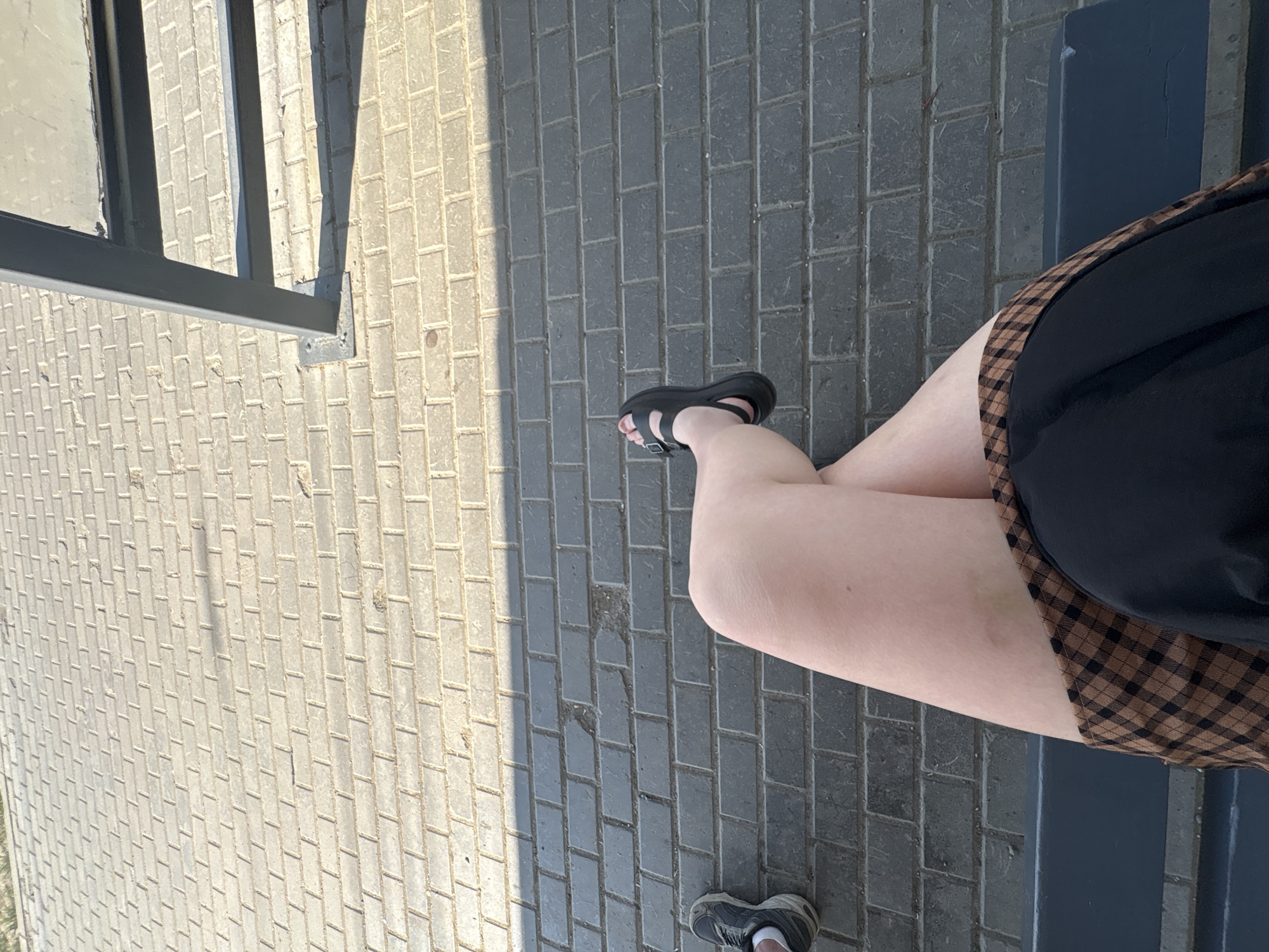 The image depicts an overhead shot of a person sitting on a bench on a paved surface. The person is wearing a black dress with a brown checkered trim, and black sandals on their feet. One leg is crossed over the other. A grey bench is visible beneath part of the person. The paving stones are grey, with sunlight casting shadows across them. A metal structure is visible in the top left corner of the image. There is no information in the image that can confirm the location of the city of Izhevsk, Russia.