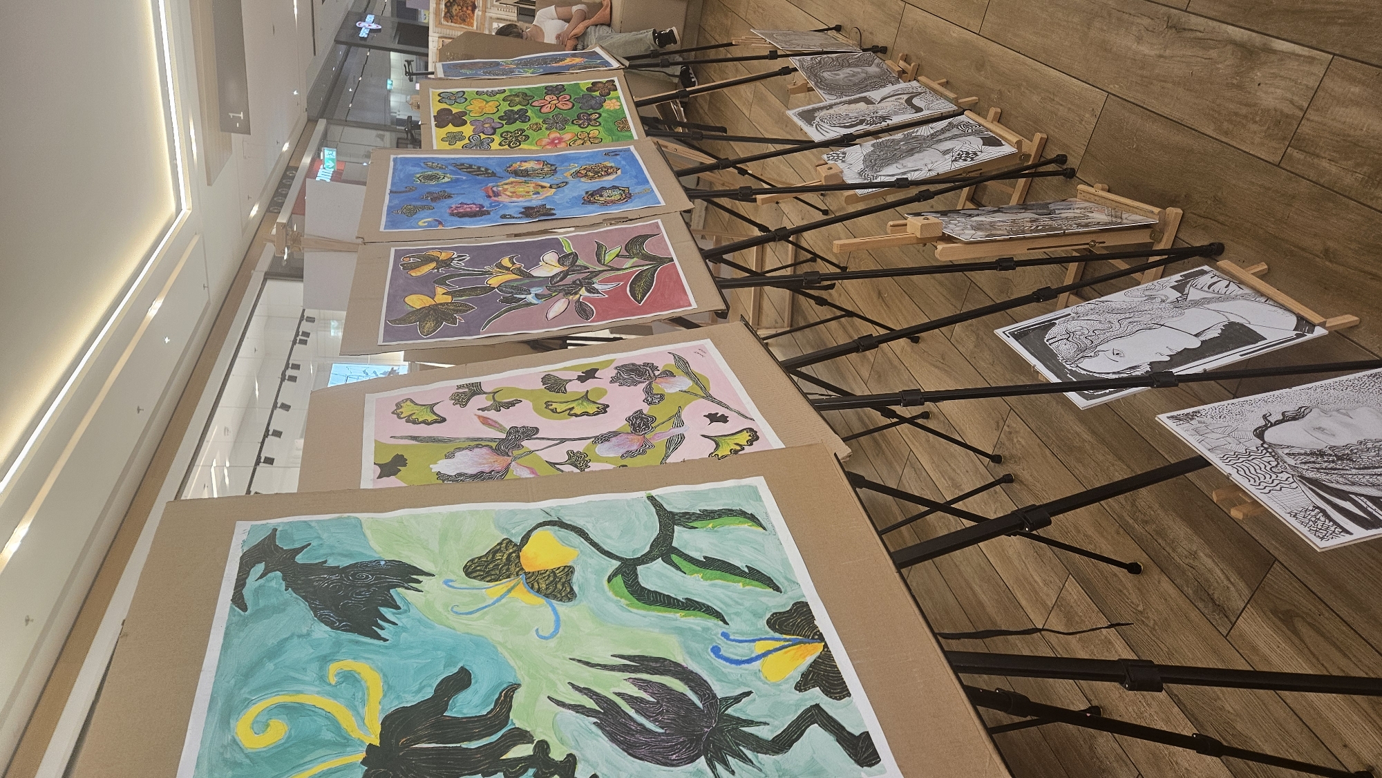 The image shows an art exhibition featuring paintings and drawings displayed on easels. The exhibition appears to be held indoors, possibly in a shopping mall, given the lighting and the visible storefronts in the background. The artworks include colorful floral and abstract pieces on larger easels, as well as black and white portrait drawings on smaller easels positioned at the front. The overall impression is one of a public display of art. The location is identified as being in the city of Prešov, Slovakia.