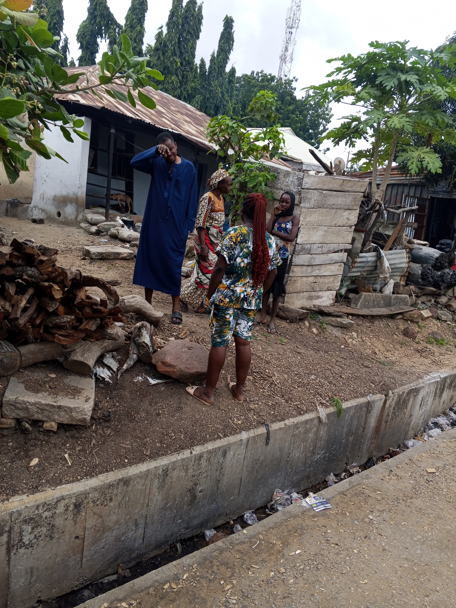 In Zango-ndaji, Nigeria, a group of four individuals stands near a makeshift dwelling with a corrugated metal roof and rough wooden walls. A man in a blue robe is positioned on the left, covering his eyes with his hand. To his right, an elderly woman wears a patterned headwrap and dress. Further to the right, two younger women are present. One wears a vibrant, patterned outfit consisting of a top and shorts, her hair in long, reddish braids. The other young woman is in a dark floral-patterned dress and a black headscarf. The ground is a mixture of dirt, gravel, and scattered debris, including a pile of chopped wood, rocks, and discarded items. A concrete drainage ditch runs along the foreground, containing stagnant water and litter. In the background, tall trees and a telecommunications tower are visible against an overcast sky.
