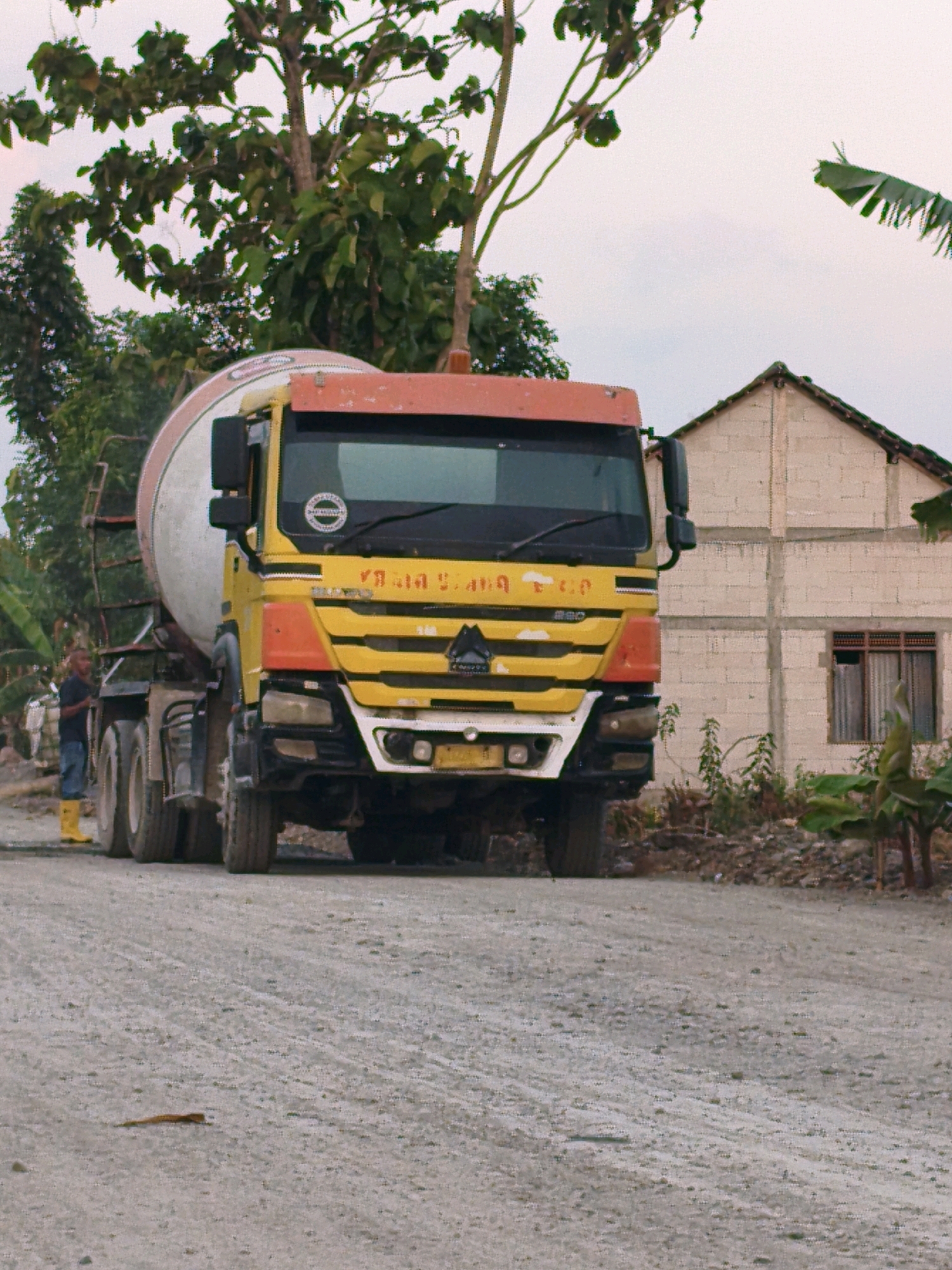 A yellow and orange cement mixer truck is parked on a dirt road. A man wearing blue jeans and yellow boots stands near the front of the truck. In the background, there is a beige, unfinished building with a brown window and a tiled roof. Green foliage and trees are visible around the building and behind the truck. The sky is overcast. The scene appears to be in a rural or semi-rural area in Gondangrejo, Indonesia.