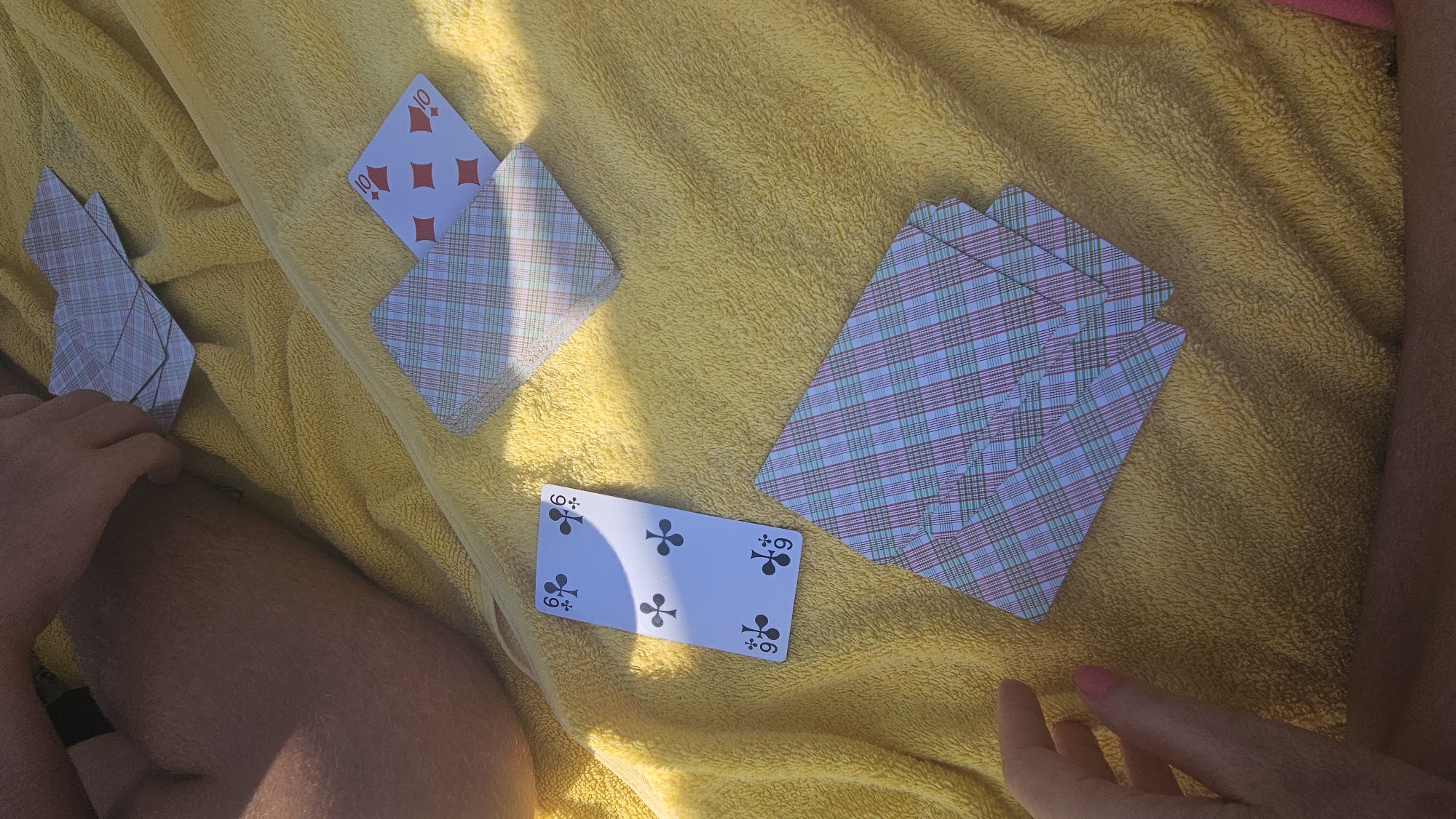 In L'Île-Rousse, France, two individuals are engaged in a card game on a yellow towel. One person's legs and hand are visible, with their hand positioned over a stack of cards. Scattered across the towel are several playing cards. One card, a six of clubs, is clearly displayed, along with a ten of diamonds. The remaining cards have their backs facing upwards, showing a plaid pattern. The scene is illuminated by sunlight, casting shadows on the towel.