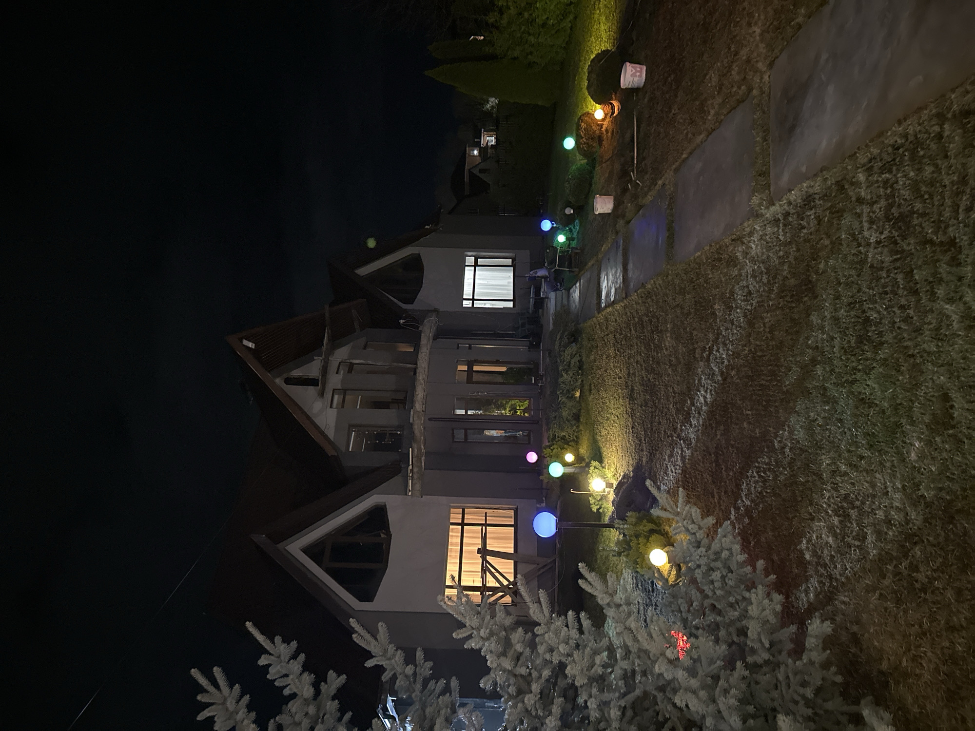 The image depicts a house at night, possibly in the city of Kursk, Russia. The house has a gabled roof and appears to be under construction, evidenced by the scaffolding visible in one of the windows. The house is lit from within, and the exterior is decorated with spherical lights along a path leading to the house. There are white trees or bushes in the foreground and greenery around the path. The sky is dark.