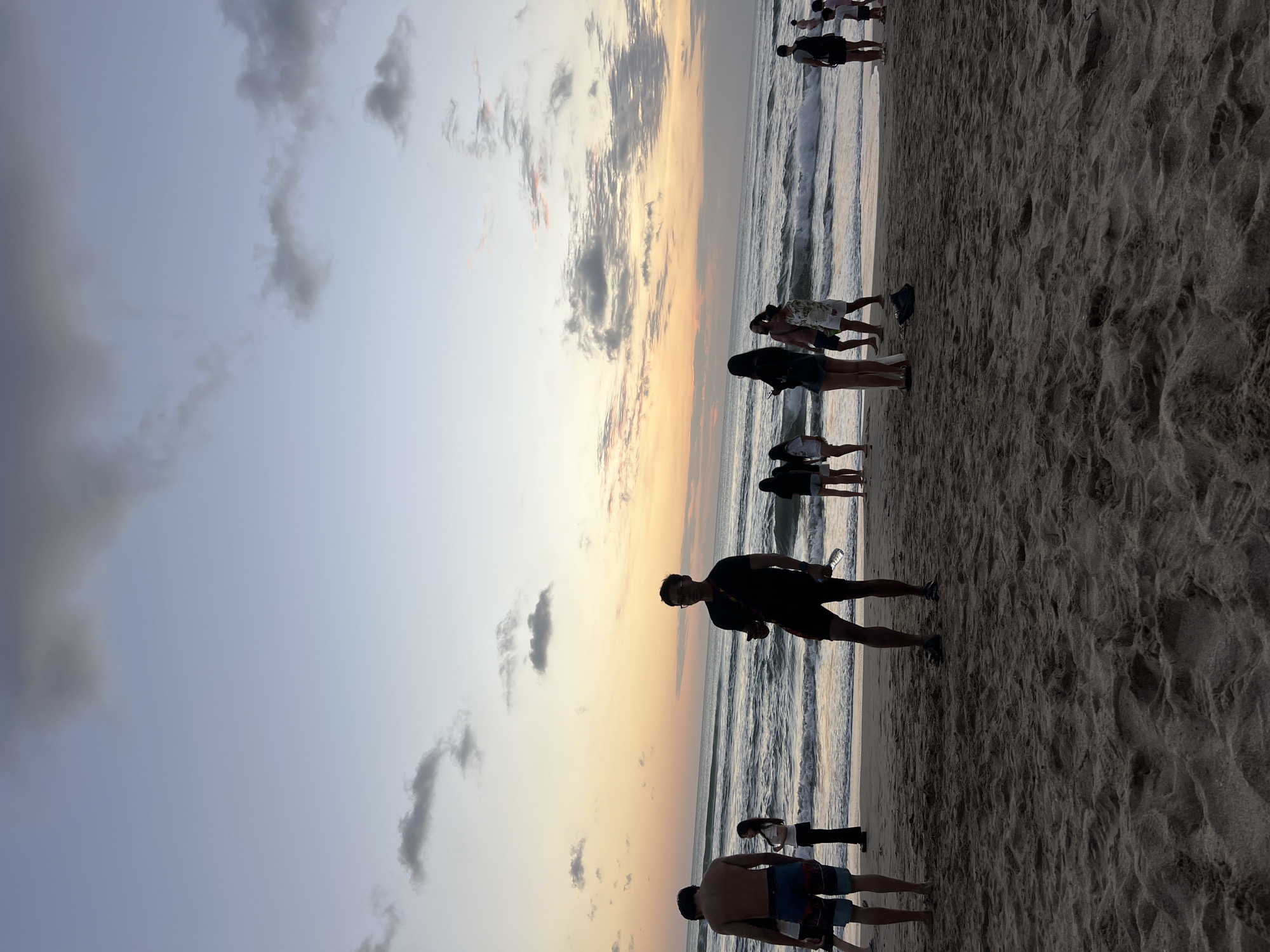 The image depicts a sunset scene on Kuta Beach, Indonesia. Several individuals are present on the sandy shore, facing the ocean. The sky is filled with muted colors of orange, yellow, and blue, with scattered clouds. The waves are gently rolling onto the beach, creating a serene atmosphere. Some people are walking, while others are standing and observing the sunset. The overall impression is one of a relaxed evening at a popular tourist destination.