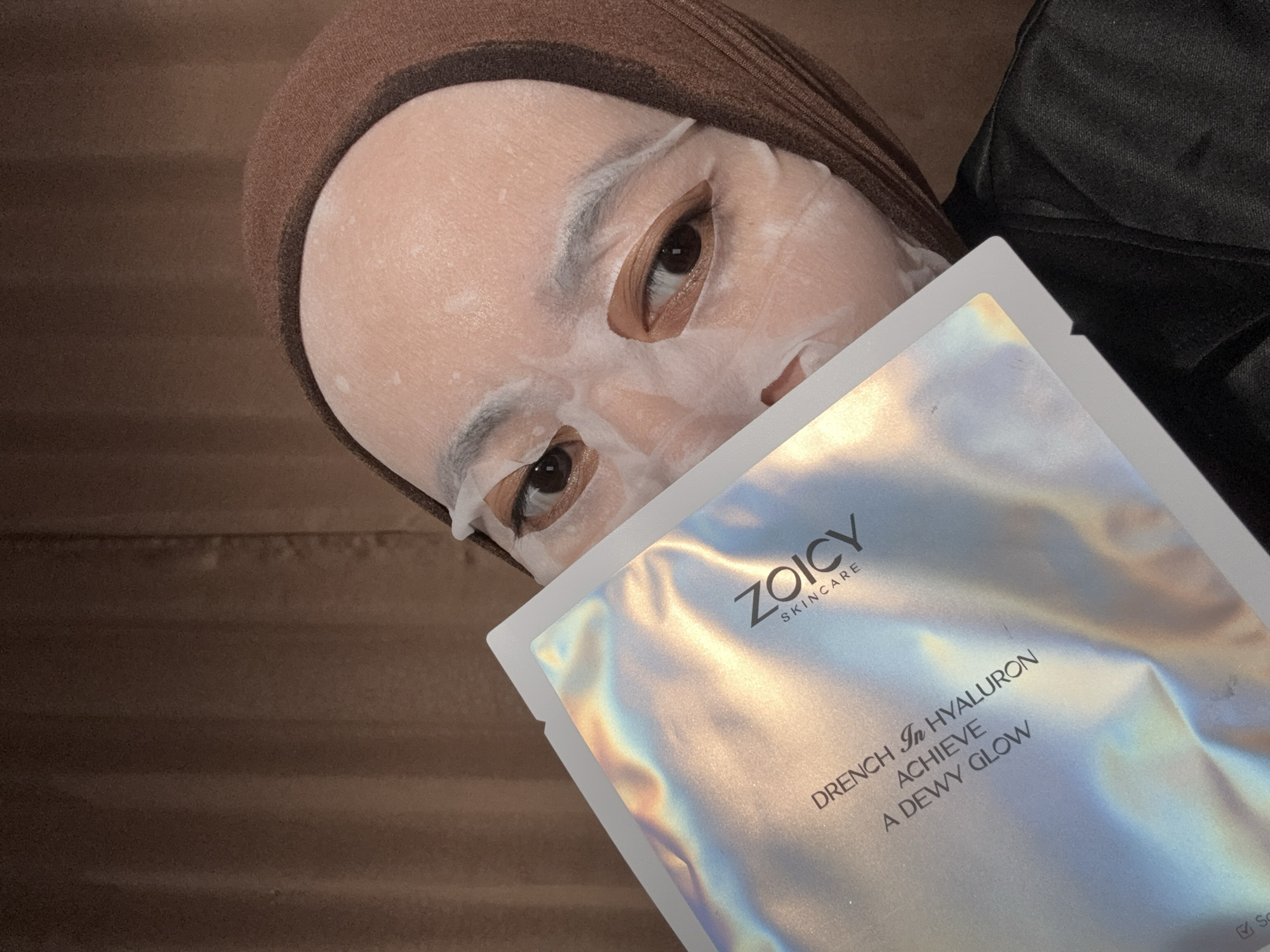 The image captures an intimate, angled close-up of a person engaged in a skincare routine indoors. The subject, likely female, wears a white sheet mask on their face, with cutouts allowing their dark eyes to be visible, framed by light-colored eyelids. A brown head covering, possibly a hijab or turban, frames their face, and they appear to be wearing a dark garment. Their expression seems neutral and calm.
They are holding an iridescent, silver-holographic sheet mask packet from "ZOICY SKINCARE," partially covering the lower portion of their face. The packet clearly displays the brand logo and the text: "DRENCH In HYALURON" and "ACHIEVE A DEWY GLOW," indicating the product's purpose. The background consists of a soft, dark brown surface, possibly a wall or upholstered furniture, suggesting a cozy and private indoor setting, consistent with an evening or nighttime skincare ritual. There are no specific cues within the image to identify the Gadingrejo, Indonesia location, other than the provided context.