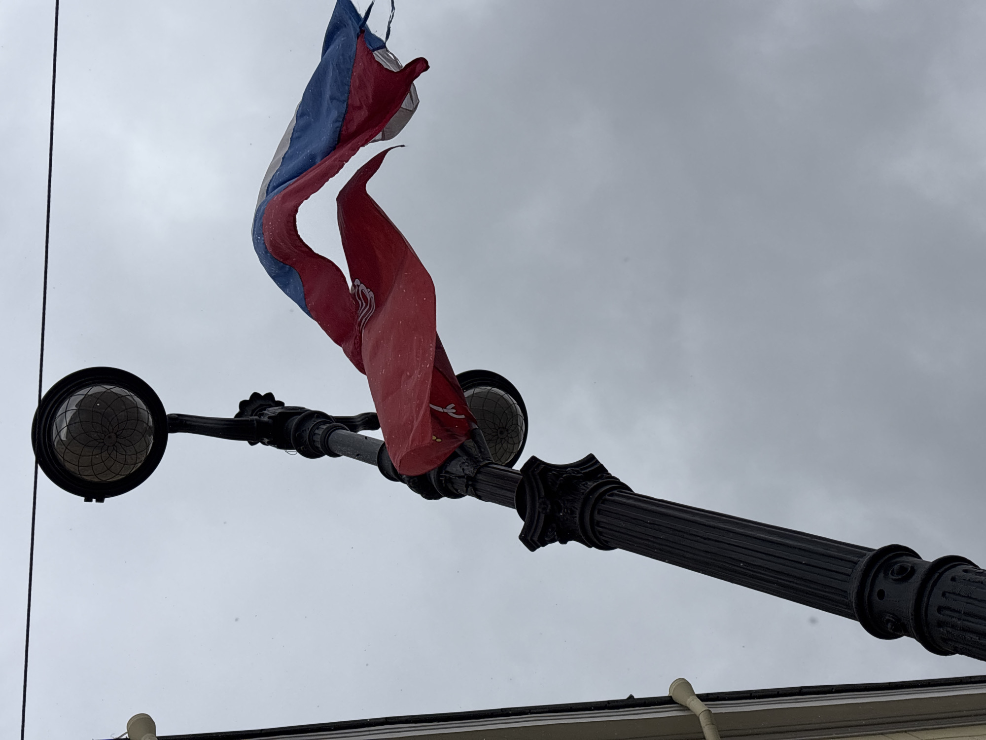 The image shows a flag of Saint Petersburg, Russia, attached to a black lamppost against a cloudy gray sky. The flag has horizontal stripes of red, blue, and white, and it is billowing in the wind. The lamppost has two globe-shaped lights on either side of the pole. The base of the lamppost is visible at the bottom of the image, attached to a building.