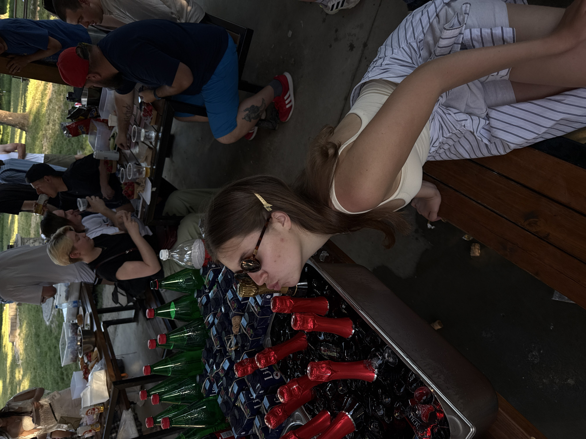 In Cukarica Urban Municipality, Serbia, an outdoor gathering unfolds at picnic tables. A woman in a light-colored tank top and striped shorts leans over a metal container filled with ice and bottles of beer and champagne; she wears sunglasses and has a hair clip. The container is densely packed with green beer bottles and red champagne bottles. Several individuals are standing or seated around the picnic tables, some interacting with food and drinks. A man in a blue shirt and red cap leans over the table. A woman with blonde hair and a black top is visible. The ground is partially visible, suggesting an outdoor setting with potential greenery nearby.