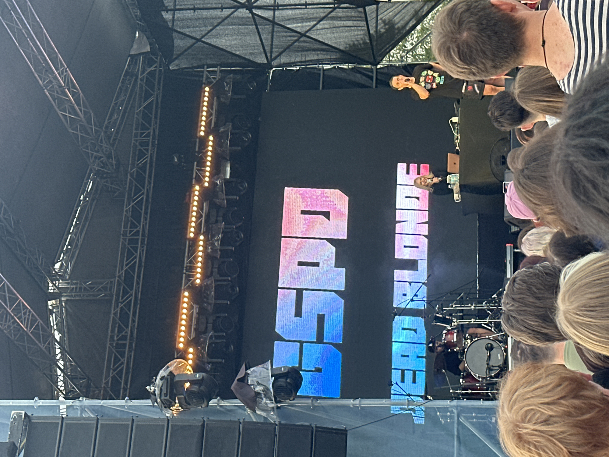 The image shows an outdoor concert stage with a large LED screen displaying the text "GDSPD" and "HEAD BLONDE". A drummer is visible on the left side of the stage, playing a drum kit. In the center, a woman is standing behind a DJ booth with a laptop and microphone, and a man is on the right side of the stage, also holding a microphone and speaking. Several spotlights are mounted above the stage. In the foreground, the backs of many people's heads are visible, indicating they are part of an audience. The concert appears to be taking place in Saint Petersburg, Russia, given the context of previous images provided. The environment suggests a festival or large outdoor event.