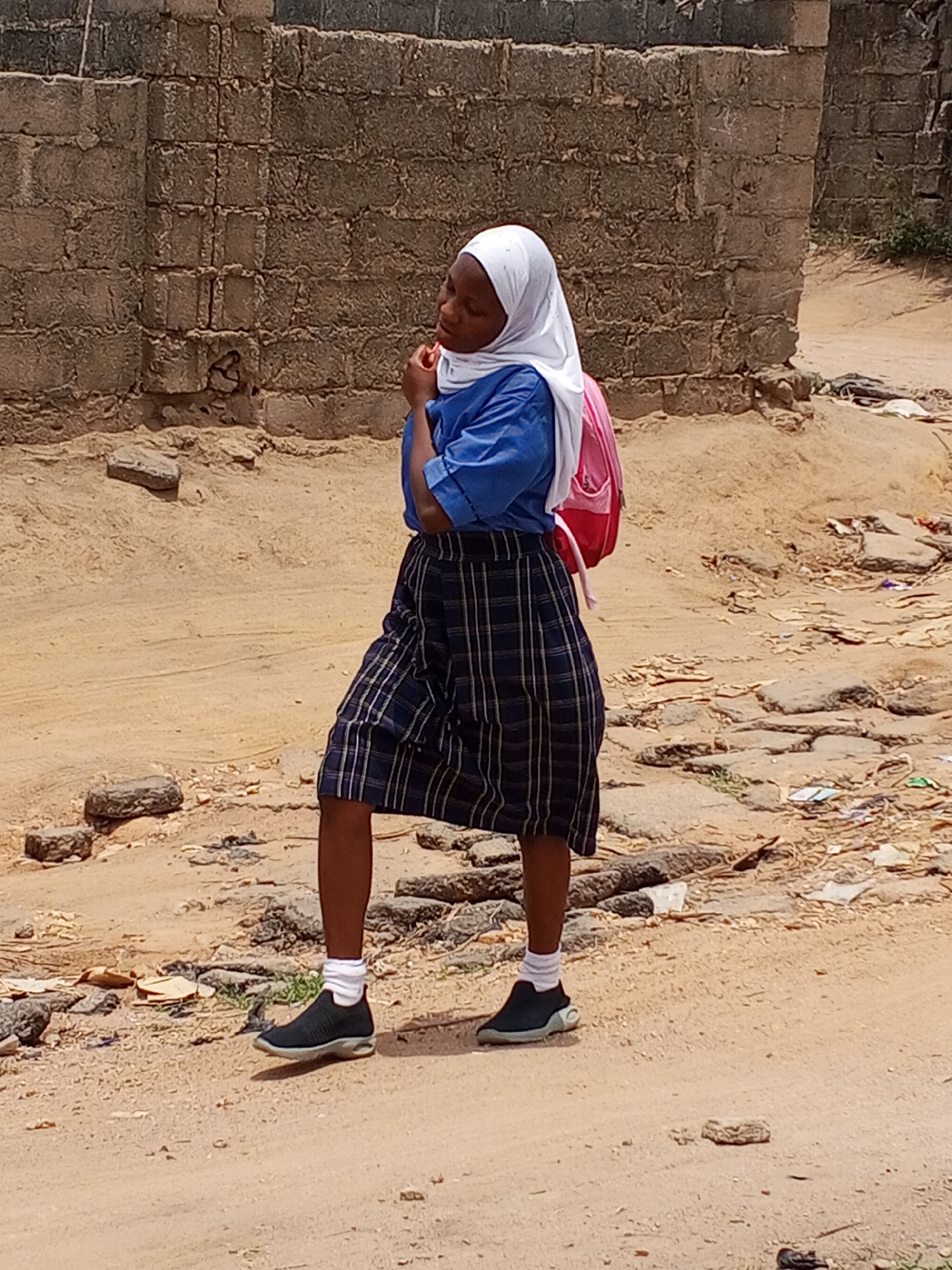 A young person in a blue collared shirt and a blue and white plaid skirt stands on a sandy and littered street, her left foot slightly raised. She wears a white headscarf and a pink backpack. She has a hand to her chin, appearing to be deep in thought. The background shows a brick wall, and the setting appears to be Adonkolo, Nigeria, judging by the style of construction and surrounding environment.