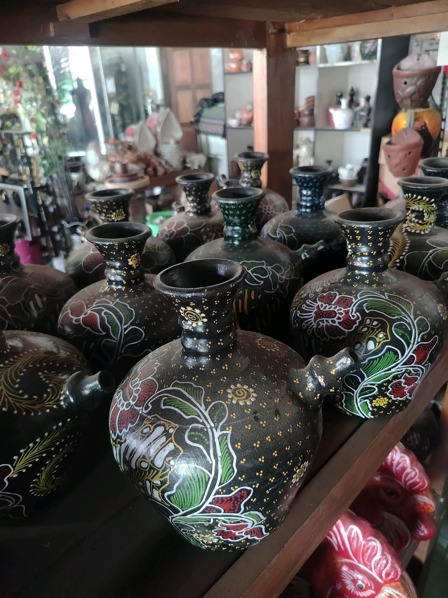 A shelf in a shop in Kroya, Indonesia, displays several traditional Indonesian ceramic jugs with intricate batik-inspired patterns. The jugs are primarily black with vibrant floral motifs in red, green, and white, accented by small yellow dots. They have narrow necks and curved handles. In the background, shelves are filled with various pottery items, including what appear to be decorative vases and planters. In the foreground, the heads of several decorative red ceramic roosters are visible on a lower shelf. The lighting is somewhat dim, creating shadows that highlight the textures and designs of the ceramics.