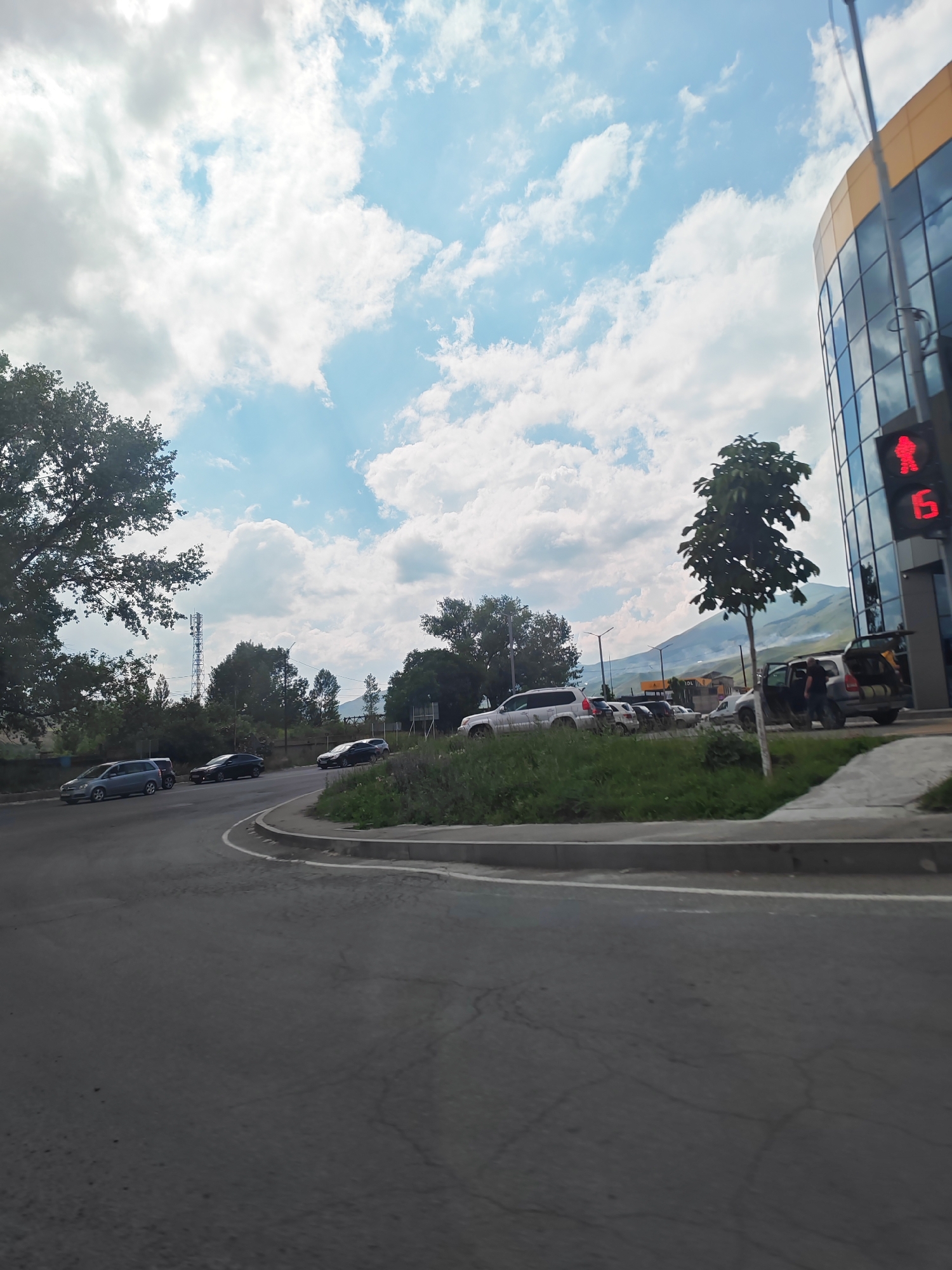 The image shows a street scene in Vanadzor, Armenia, with a focus on cars, a building, and the sky. In the foreground, a paved road curves to the left. Several cars are visible parked and driving on the road. A light-colored SUV is parked on the right side of the road, with its rear hatch open. A person in a dark shirt is standing next to it. Further down the road, a modern building with a glass facade and a yellow-tinted top is prominent on the right. A pedestrian traffic light showing a red silhouette of a person is mounted on a pole in front of the building. The sky above is filled with fluffy white clouds against a bright blue background. In the distance, a cellular tower stands tall amongst trees, and a hazy mountain range is visible. The environment appears to be an urban or semi-urban area with elements of nature.