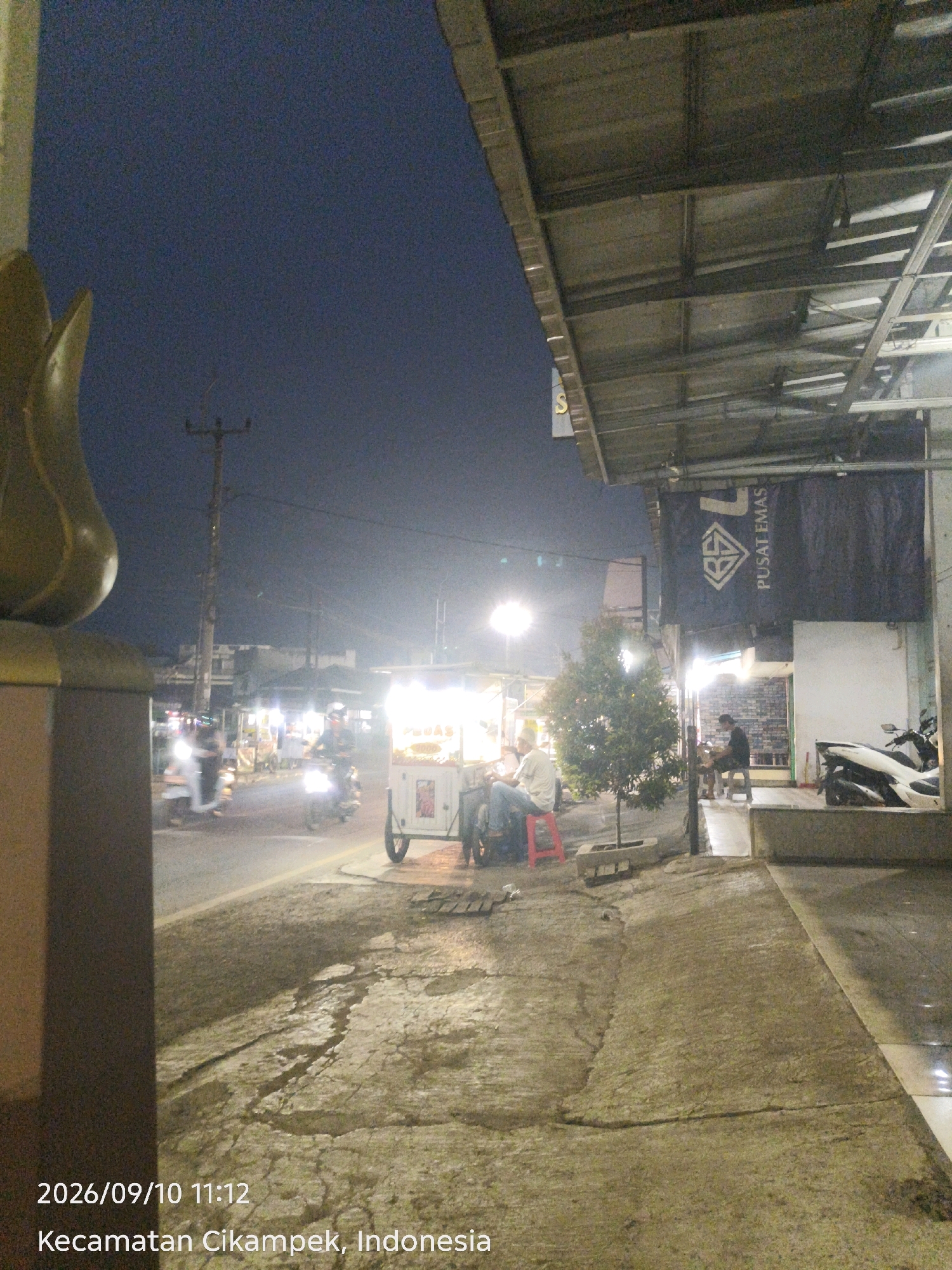 A street-level, medium shot captures a nighttime scene in Cikampek, Indonesia. The sky is dark blue, indicative of late evening or early morning. In the foreground, a textured, cracked concrete sidewalk runs along the right side of the image, leading to a paved area with some reflective surfaces. A substantial, dark brown, pillar-like structure is visible on the far left, partially framing the scene.
The street itself is paved and shows signs of wetness, with reflections of lights. Several motorcycles are present: one is parked on the sidewalk, and others are in motion on the street, their headlights illuminated. A food cart with bright lights and a white exterior is positioned on the sidewalk, with people seated on stools nearby. One person, wearing a light-colored shirt, is seated on a red stool, looking away from the camera. Another individual, in darker clothing, sits on a stool further back, near a tree.
Above the food cart and the area with seating, a covered structure with a corrugated metal roof and exposed beams extends over the sidewalk. A dark banner with white text and a diamond logo is displayed on this structure, with the text "PUSAT EMAS" visible. A small, leafy tree is planted in a concrete planter box on the sidewalk. In the background, buildings and streetlights are visible, contributing to the overall ambient lighting. The timestamp "2026/09/10 11:12" and the location "Kecamatan Cikampek, Indonesia" are overlaid on the bottom left of the image.