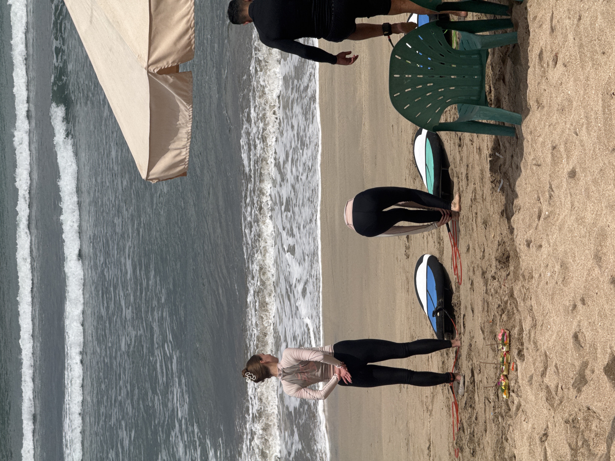 An outdoor scene on a sandy beach in Kuta, Indonesia, shows several people and surfboards near the ocean. In the foreground, a green plastic chair sits on the sand. To the left of the chair, a surfboard with a blue and white design is propped up. Nearby, several small offerings with incense sticks are placed on the sand.
A woman in a light pink long-sleeved rash guard and black leggings stands with her back to the camera, facing the ocean. Her hands are clasped behind her back, and her hair is styled in a bun.
In the mid-ground, another person, also wearing black leggings and a light-colored rash guard, is bent over, seemingly adjusting their attire or a surfboard. Two surfboards are visible on the sand in front of this individual.
To the right, a man in a black long-sleeved rash guard and black shorts is walking away from the camera, towards the right edge of the frame. His left ankle is visible, and he appears to be wearing a leash for a surfboard.
The ocean in the background exhibits moderate waves with white foam breaking near the shore. The sky is overcast, casting a diffused light on the scene. An open umbrella with a light tan canopy is visible in the upper right portion of the image. The overall impression is of a group preparing for or finishing a surfing activity on a typical day in Kuta.
