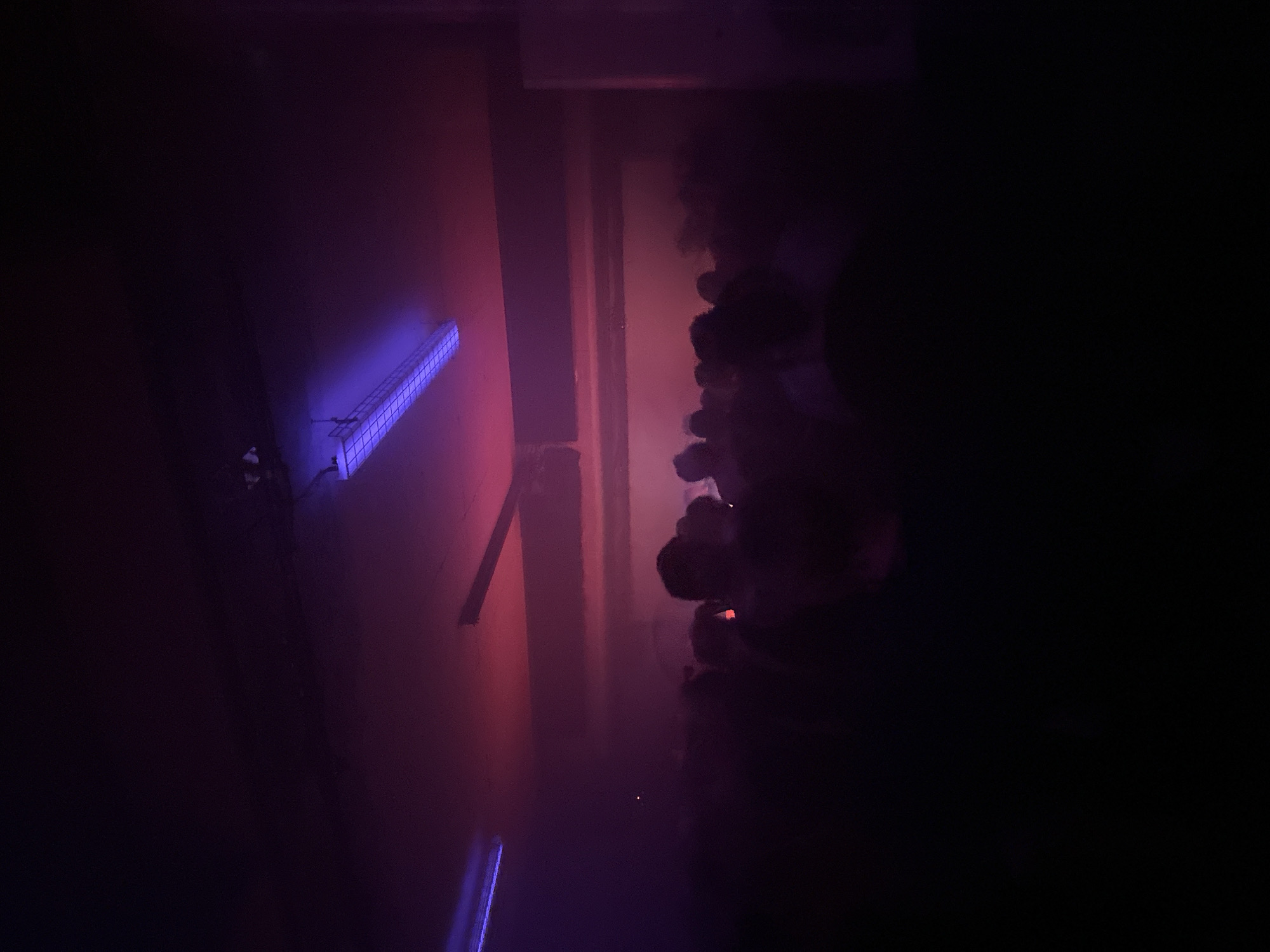 The image depicts a dark, enclosed space with a mix of blue and red lighting. On the left side, a blue fluorescent light fixture is mounted on a wall with a reddish hue. There appears to be a doorframe or wall separating a dimly lit area with a possible crowd of people. Some figures appear to be holding objects, possibly cigarettes, as small red glows are visible. The scene is obscured by darkness, with details difficult to discern, but the overall impression is that of an indoor event or gathering in low light conditions. There is no indication of Paris, France.