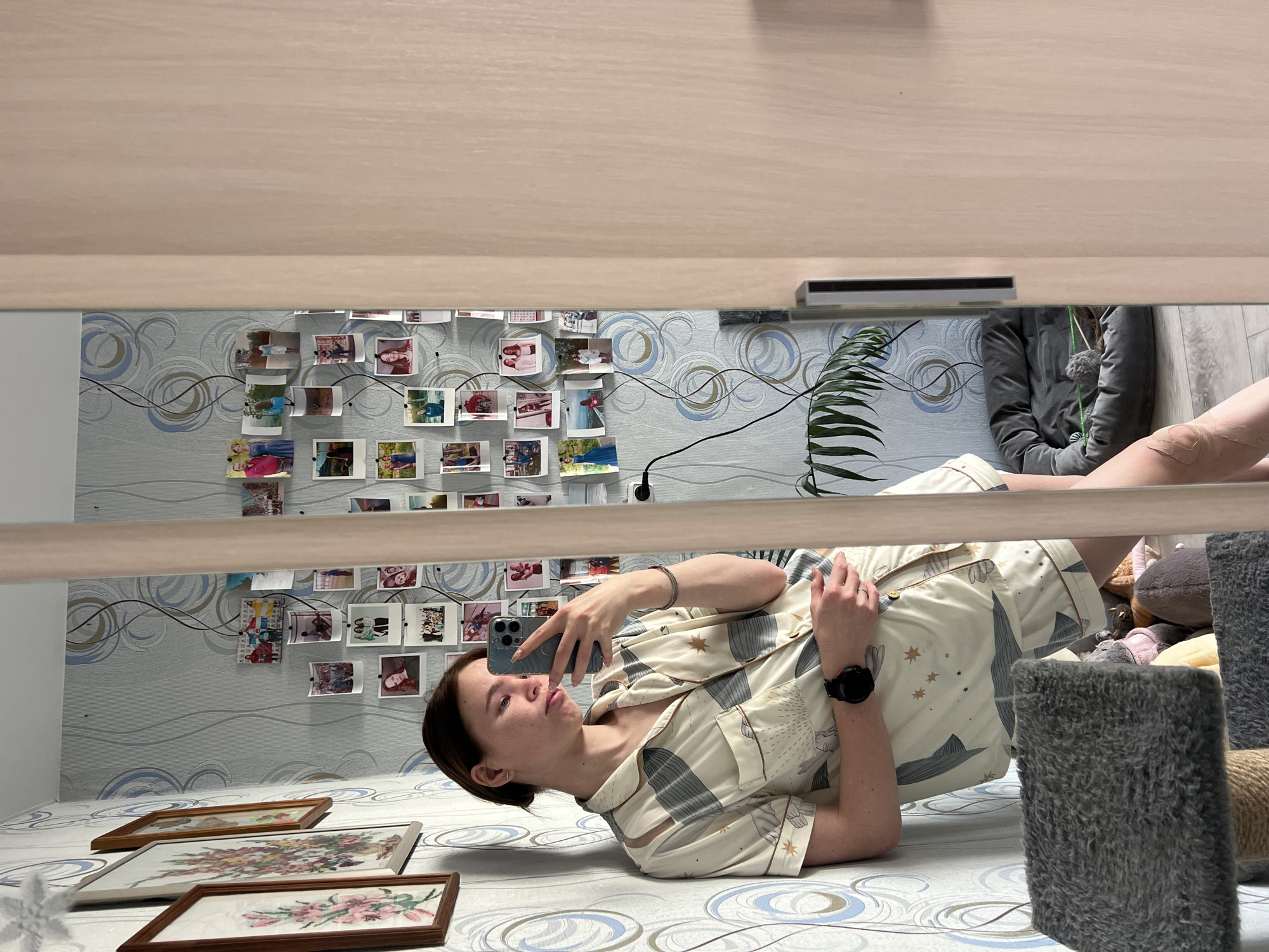 A reflected image shows a young woman lying on a bed, taking a photo of herself with her phone. She has dark hair pulled back and is wearing patterned pajamas and a smartwatch. Behind her, a wall is decorated with patterned wallpaper and a string of pinned-up photographs. Three framed embroidery pieces are positioned at the foot of the bed. A potted plant and a person seated in a gray hooded jacket can also be seen in the reflection. The city and country in the prompt (Chaikovsky, Russia) are not verifiable.