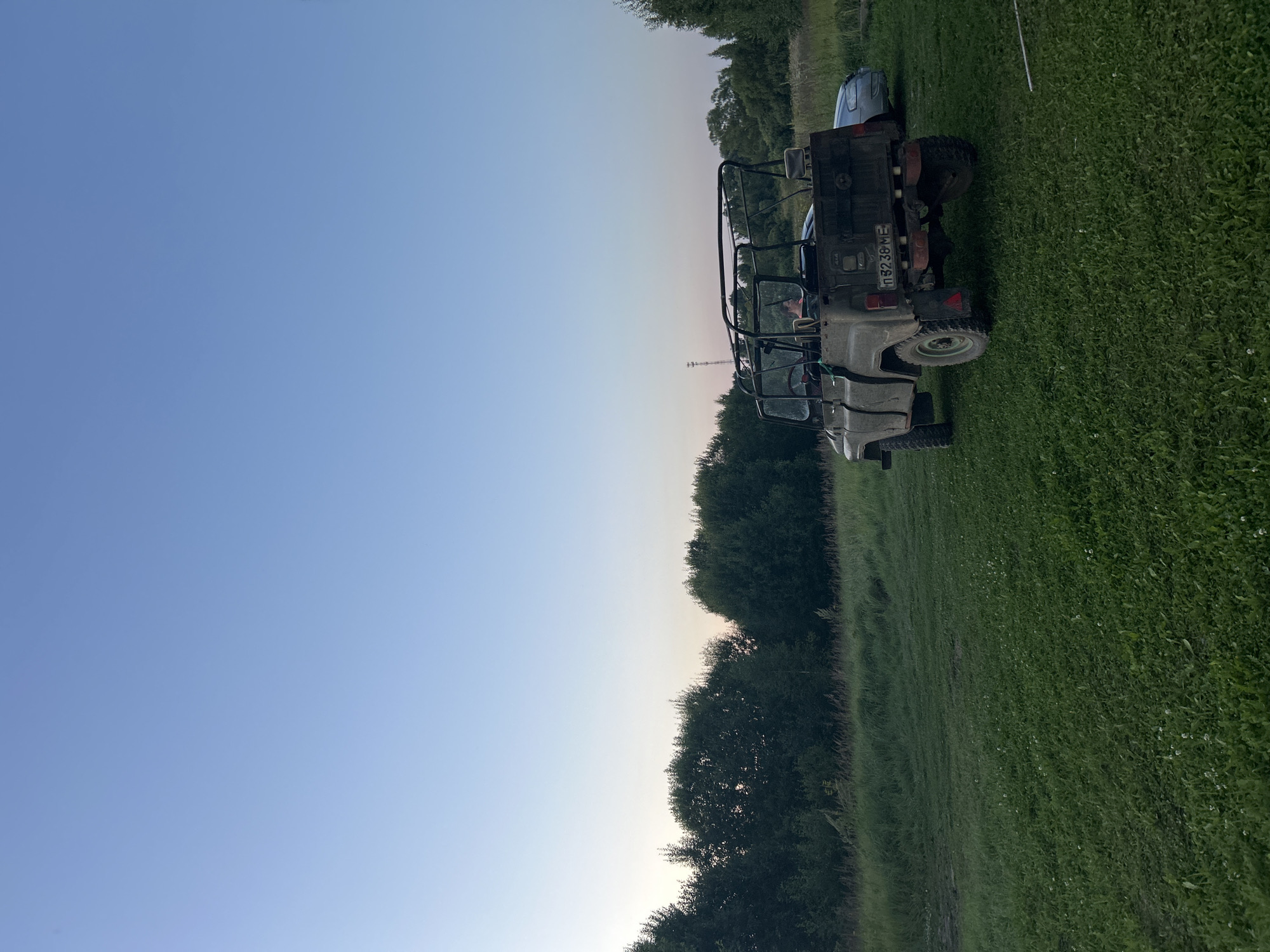 An off-road vehicle is parked on a grassy field at dusk, with a line of trees in the background. A person is visible in the driver's seat of the vehicle. A second, lighter-colored vehicle is parked to the right of the off-road vehicle, partially obscured. The sky is a clear, pale blue, transitioning to a lighter hue near the horizon. The overall scene depicts an outdoor rural setting.