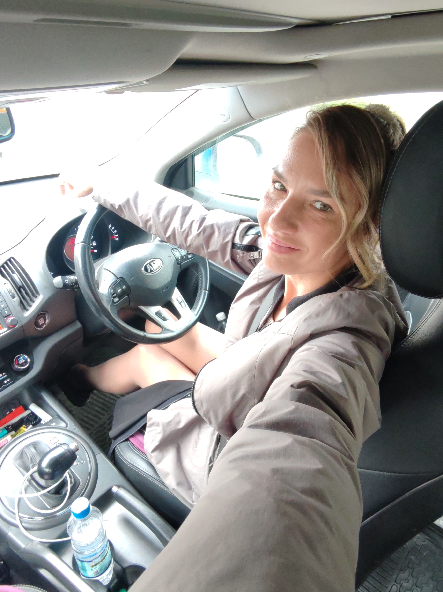 In the interior of a Kia Sportage, a blonde woman with light brown eyes is seated in the driver's seat, holding the steering wheel. She is wearing a light brown jacket. The woman is looking at the camera, smiling. The car's dashboard is visible, including the steering wheel, air conditioning vents, and a gear shift. A bottle of water is in the center console. The scene appears to be a street in Petropavlovsk-Kamchatsky, Russia, with overcast skies.