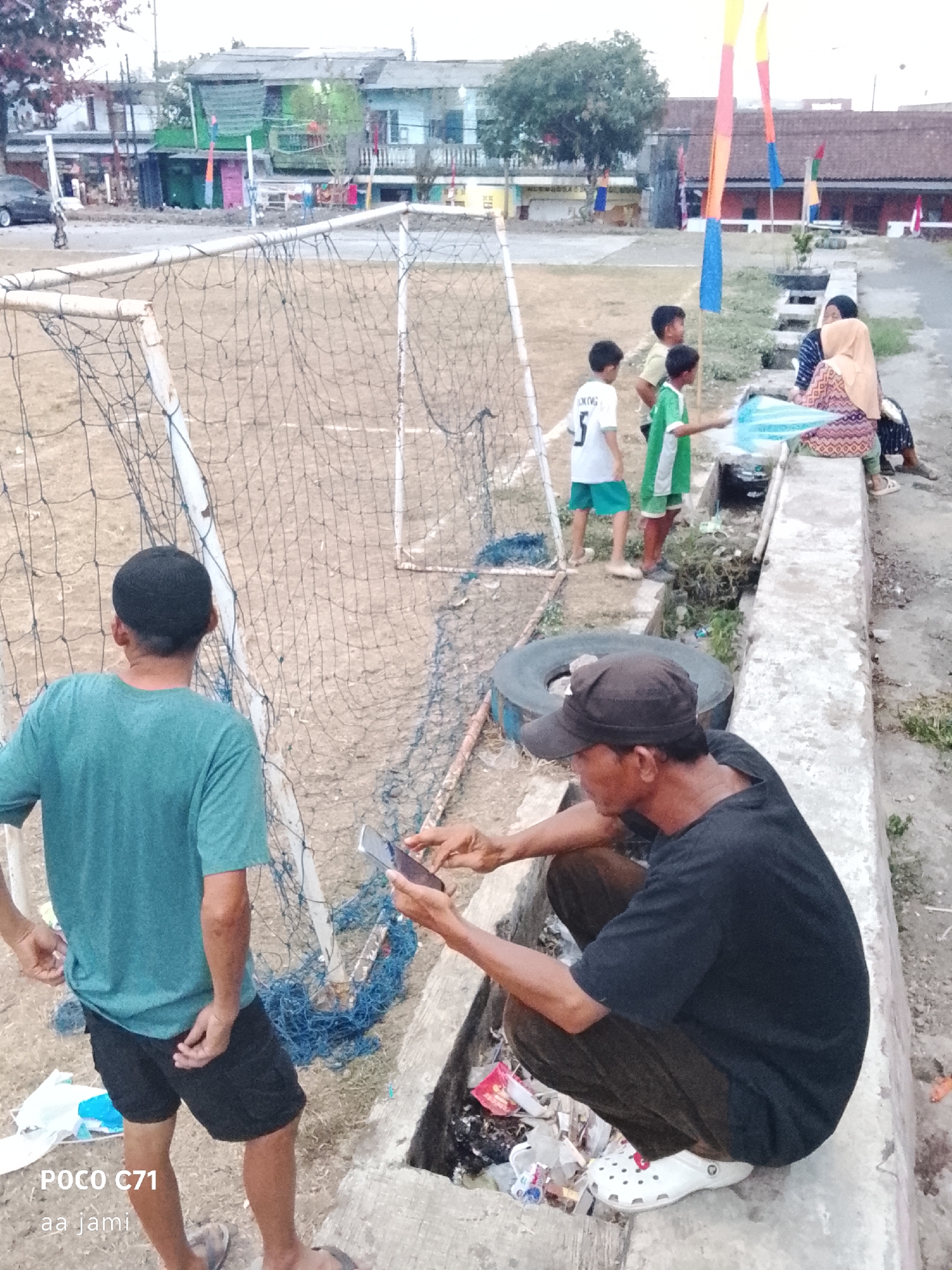 The image depicts an outdoor scene, possibly a street or a park area, in Tasikmalaya, Indonesia. In the foreground, a man is squatting and looking at his phone, wearing a black t-shirt and dark pants. Next to him, another person wearing a teal t-shirt and black shorts is standing and also appears to be engaged with a phone. A soccer net is visible to the left. Further back, several children are gathered, and two women are seated on a raised concrete structure. The background shows buildings and a street. The overall environment suggests a casual gathering in a somewhat urban setting.