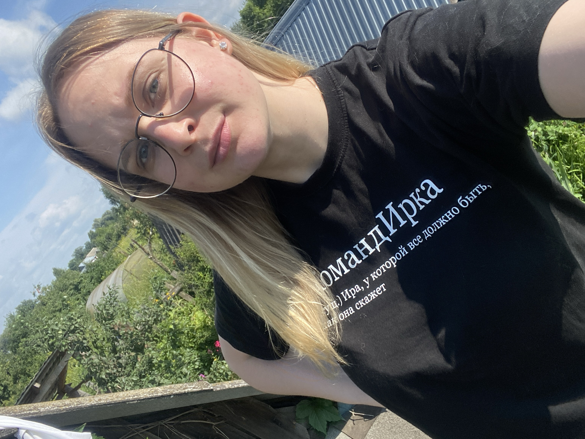 A young woman with blonde hair and glasses takes a selfie outdoors. She is wearing a black t-shirt with white text that reads "Команда Ирка, у которой все должно быть, как она скажет" which translates to "Team Ira, for whom everything must be as she says." The background shows a rural setting with greenery, a greenhouse, and a blue sky. The woman is positioned in the foreground, with the landscape behind her.