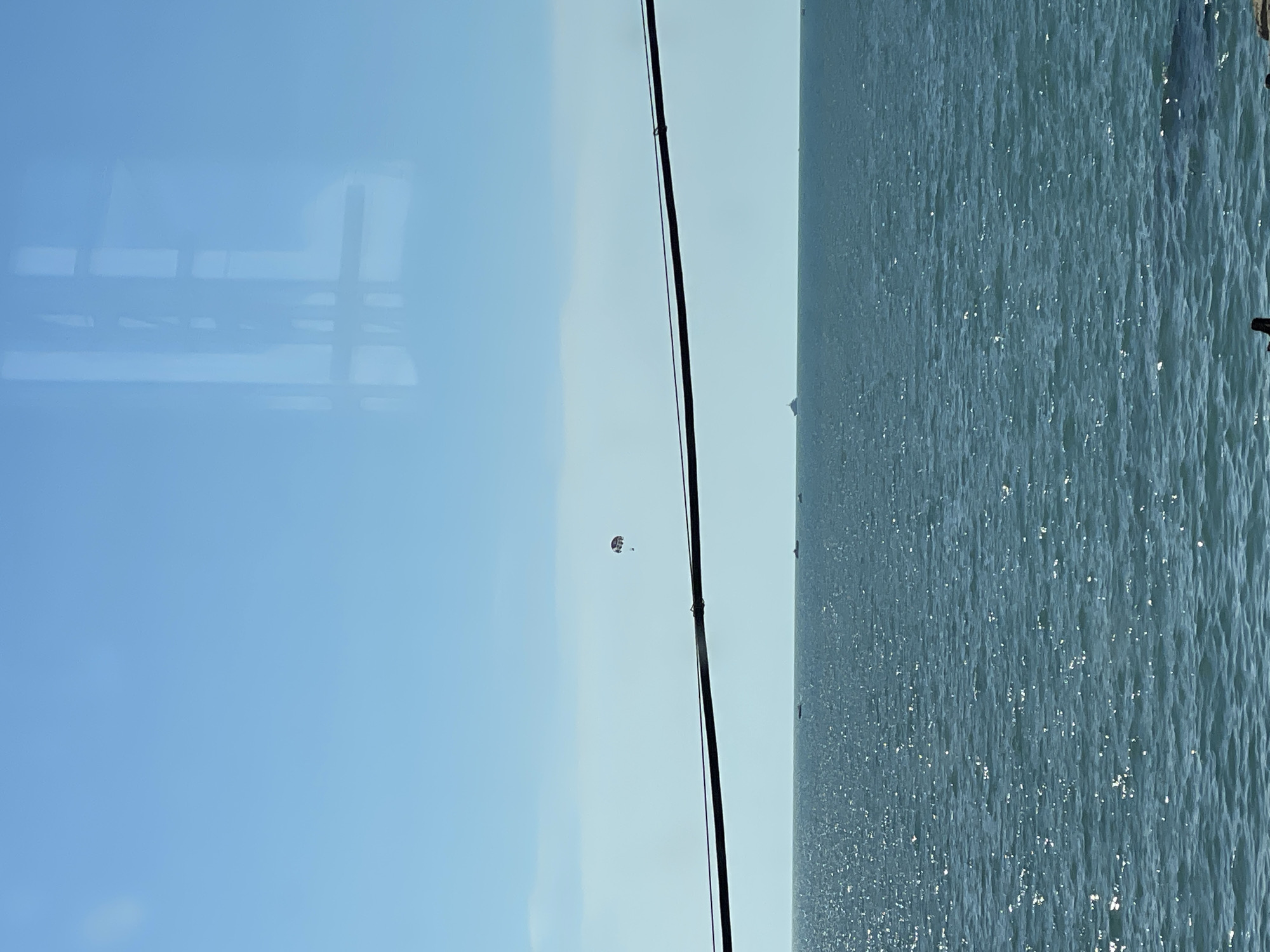 The image displays a bright, clear day with a vast expanse of calm, rippling water. The water is a deep blue and reflects the sunlight, creating a shimmering effect across its surface. In the distance, the horizon is faintly visible, and a thin, dark line suggests the presence of distant land or structures.
On the left side of the image, the sky is a pale blue, transitioning to a lighter shade towards the horizon. A faint reflection of what appears to be a window frame or structural elements is visible in the sky, suggesting the photo might have been taken from indoors or through a transparent surface. A thin, dark cable stretches vertically through the frame, possibly part of a boat or structure.
In the middle distance of the sky, a small object is visible, appearing to be a parasail with people attached. The parasail is colorful and distinct against the light sky, indicating an activity of recreational water sports is taking place.
The overall scene evokes a sense of tranquility and leisure, typical of a day spent by the sea or a large body of water. The bright sunlight suggests it is daytime, likely late morning or afternoon. The clear sky and calm water indicate pleasant weather conditions. While no specific text is visible, the context of Khostinskiy, Russia, places this scene in a coastal or lakeside resort area.