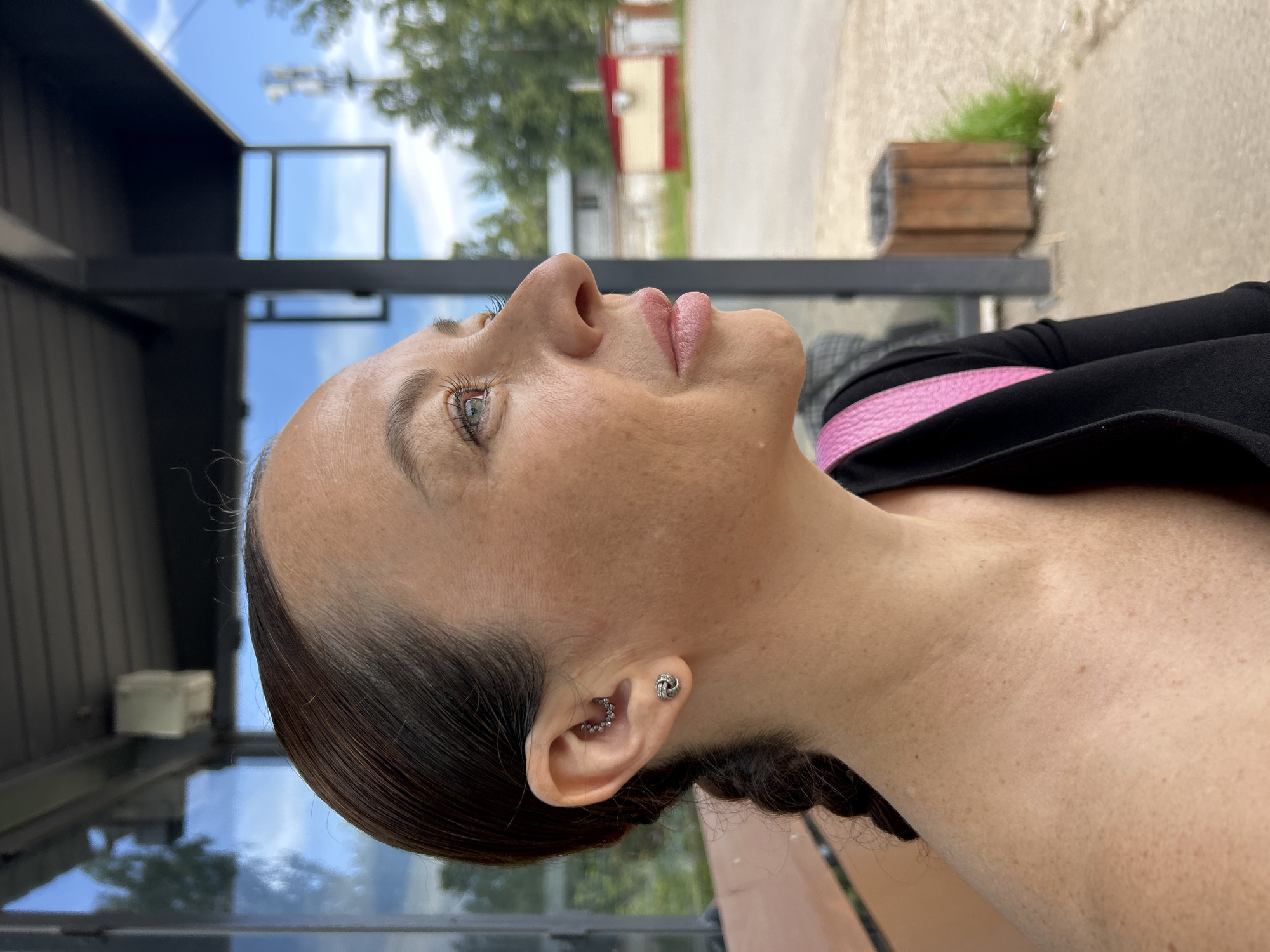 In Chaikovsky, Russia, a side profile of a woman is captured as she looks to her right. She has brown hair styled in a braid, and she is wearing a black top with a pink strap over her shoulder. Her ear has two piercings: one with a flower-shaped stud and another with a circular piece of jewelry. The background suggests an outdoor setting, possibly a covered seating area or bus stop, with a window reflecting the sky and trees. Beyond the window, there is a dirt path, a wooden trash bin, and a few buildings, including one with red and yellow accents. The lighting is bright, indicating it is daytime.
