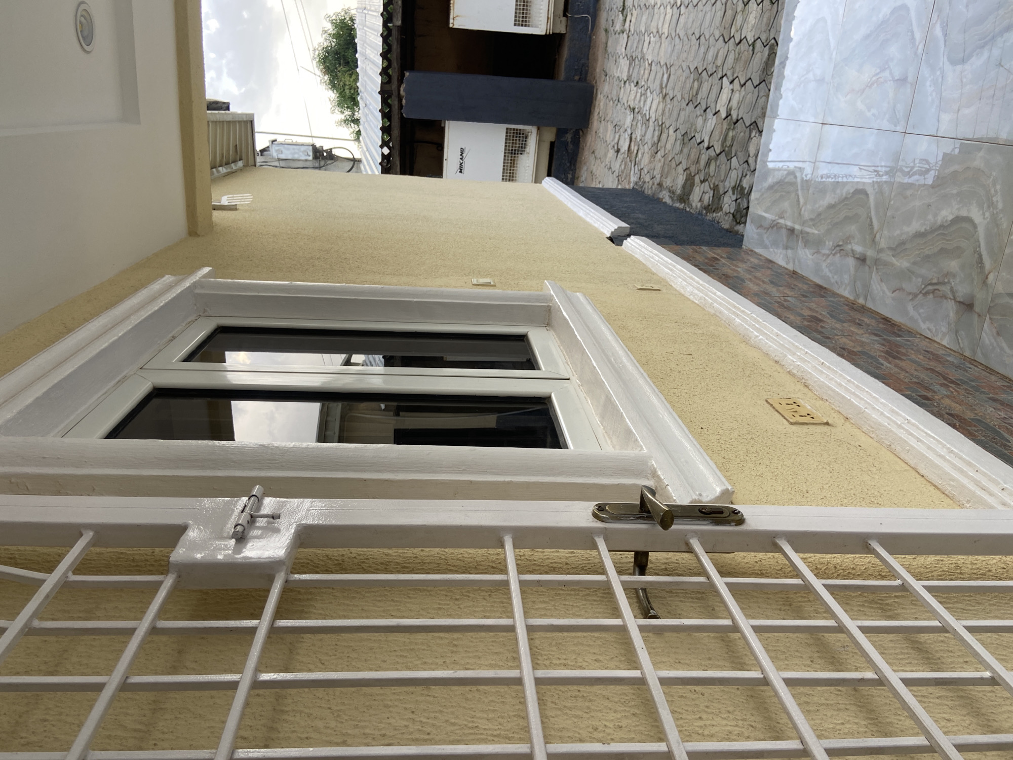 The image depicts a close-up view of a white metal security grill in front of a window on a pale yellow textured wall. The grill has horizontal and vertical bars, with a silver latch mechanism visible in the center. The window itself is white-framed and appears to have dark tinted glass, reflecting the cloudy sky.
In the background, elements of a building and its surroundings are visible. To the left, a portion of a white soffit with a recessed light fixture is seen. Beyond that, a tiled roof and what appears to be an air conditioning unit are present. To the right, a patterned tiled wall and a stone-tiled walkway suggest an outdoor area. The overall setting seems to be the exterior of a residential or commercial building, possibly in a tropical or subtropical climate given the vegetation and style of construction visible in the background.
The lighting suggests it is daytime, with overcast conditions. There is no discernible activity or people in the frame. The focus is on the architectural details of the window, grill, and wall. A small rectangular plaque with some writing is visible on the wall to the right of the window, but the text is too small to be legible. Another label, "MIKANO," is visible on an air conditioning unit in the background.