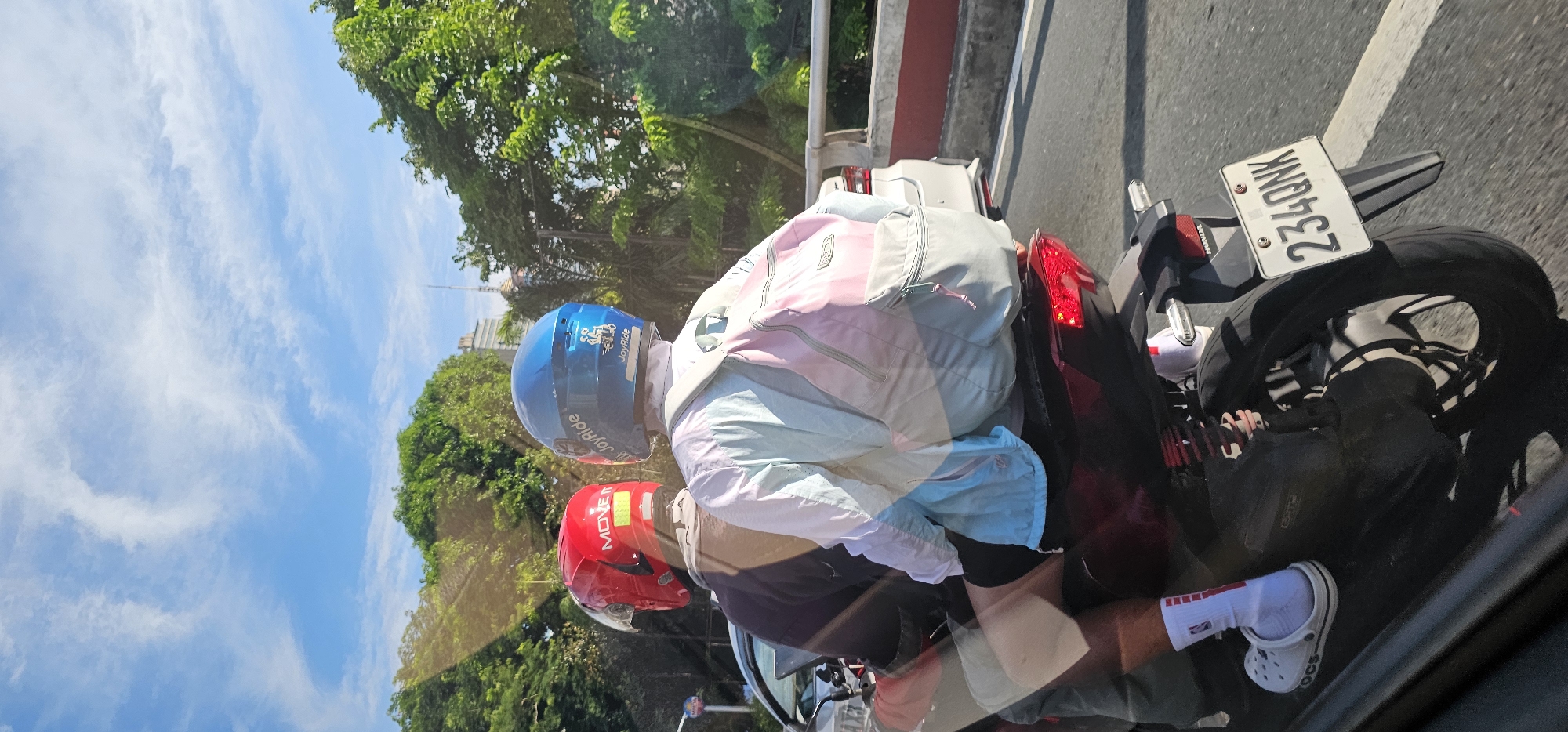 The image shows two people riding on a motorcycle, viewed from behind. The passenger in front wears a red helmet with "MOVE IT" stickers. The rider in the back wears a blue helmet with "JoyRide" logos and a light blue and pink backpack. They are on a road with trees and buildings in the background, under a blue sky with white clouds. The motorcycle has a license plate with the number "234QNK". The scene is likely in Quezon City, Philippines, as indicated by the "JoyRide" service, which is popular in the region.