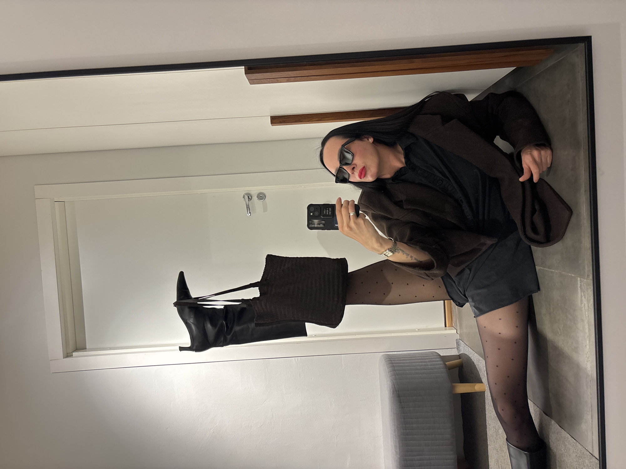 A person is captured in a mirror selfie, posing in an indoor setting. The individual has long dark hair and is wearing dark sunglasses, red lipstick, a dark brown jacket over a black shirt, black shorts, sheer black polka-dot tights, and black boots. A silver ring is visible on their right hand, which holds a black smartphone with a reflective case, pointed towards the mirror to take the photo. A silver watch is on their left wrist, and they are holding a dark brown, possibly corduroy, handbag in their left hand.
The person's left leg is raised, with the boot resting on what appears to be a white window ledge or door frame. The background reflected in the mirror shows a white door with silver handles, light grey walls, and a light grey tiled floor. A grey upholstered bench or stool is partially visible on the lower left. Wooden accents are integrated into the wall design above the door. The lighting suggests an indoor setting during the daytime. No specific text is clearly legible on the phone case or watch. While the location context is Chaikovsky, Russia, no visual cues within the image confirm this.