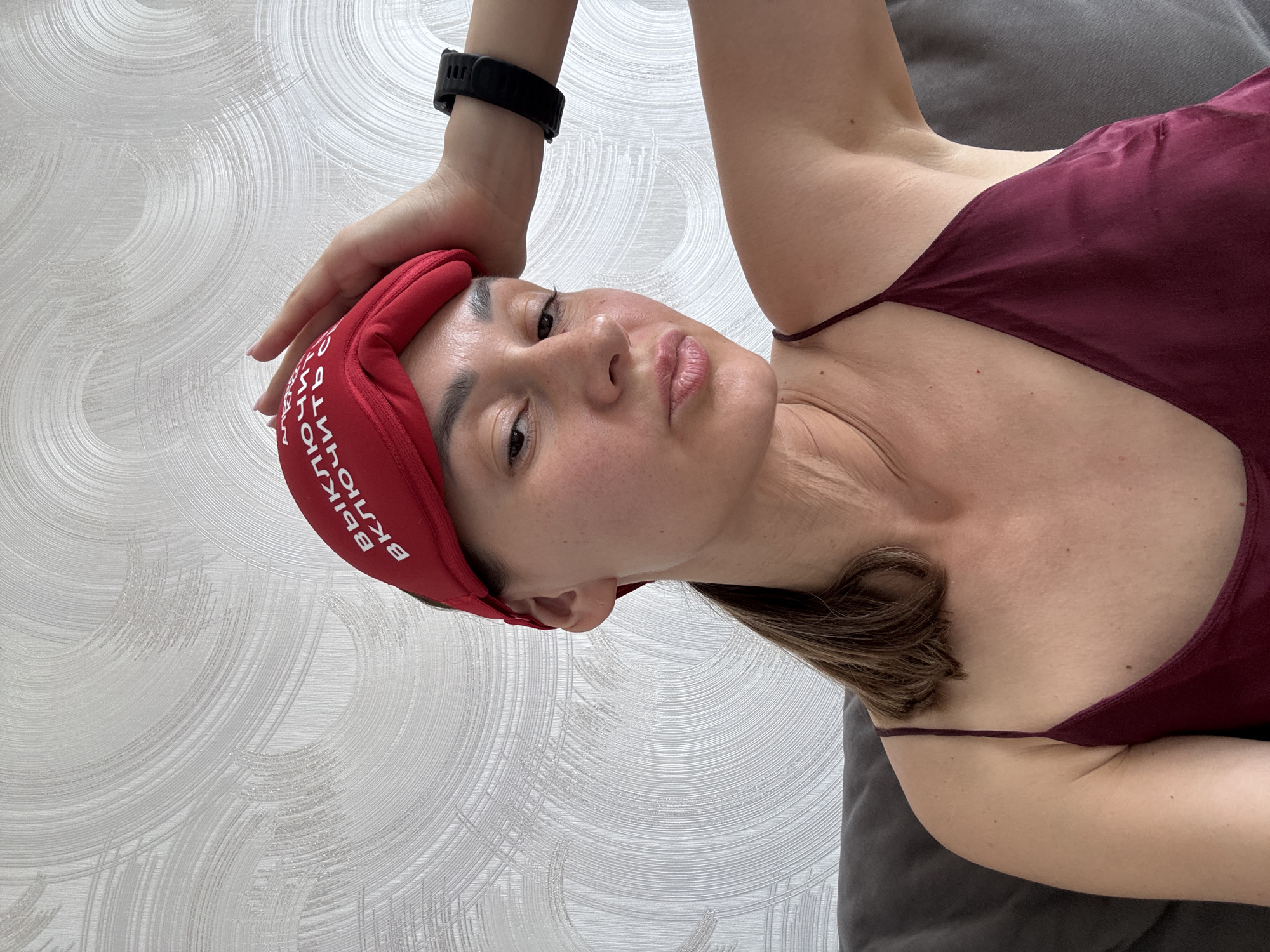 A female subject is depicted indoors in a medium-close shot, situated in Sochi, Russia. She has light-to-medium brown hair, partially visible in a ponytail. A red fabric eye mask is pushed up onto her forehead, displaying white Cyrillic text that reads "ВЫКЛЮЧИТЬ ВКЛЮЧИТЬ СЕБЯ" (transliterated as "VYKLYUCHIT' VKLYUCHIT' SEBYA"), meaning "TURN OFF / TURN ON YOURSELF." She is wearing a maroon or burgundy spaghetti strap top, possibly silk or satin. A black digital sports watch with multiple buttons is visible on her left wrist. Her left hand rests on the red eye mask, with manicured nails visible. The subject is looking directly at the camera with a playful expression, characterized by puckered lips. She is seated on a dark gray upholstered couch, partially visible in the lower left and right of the frame. The background consists of light gray or off-white wallpaper featuring an abstract, swirling, textured pattern resembling brushed metallic arcs. Lighting is even and ambient, suggesting an indoor daytime setting.