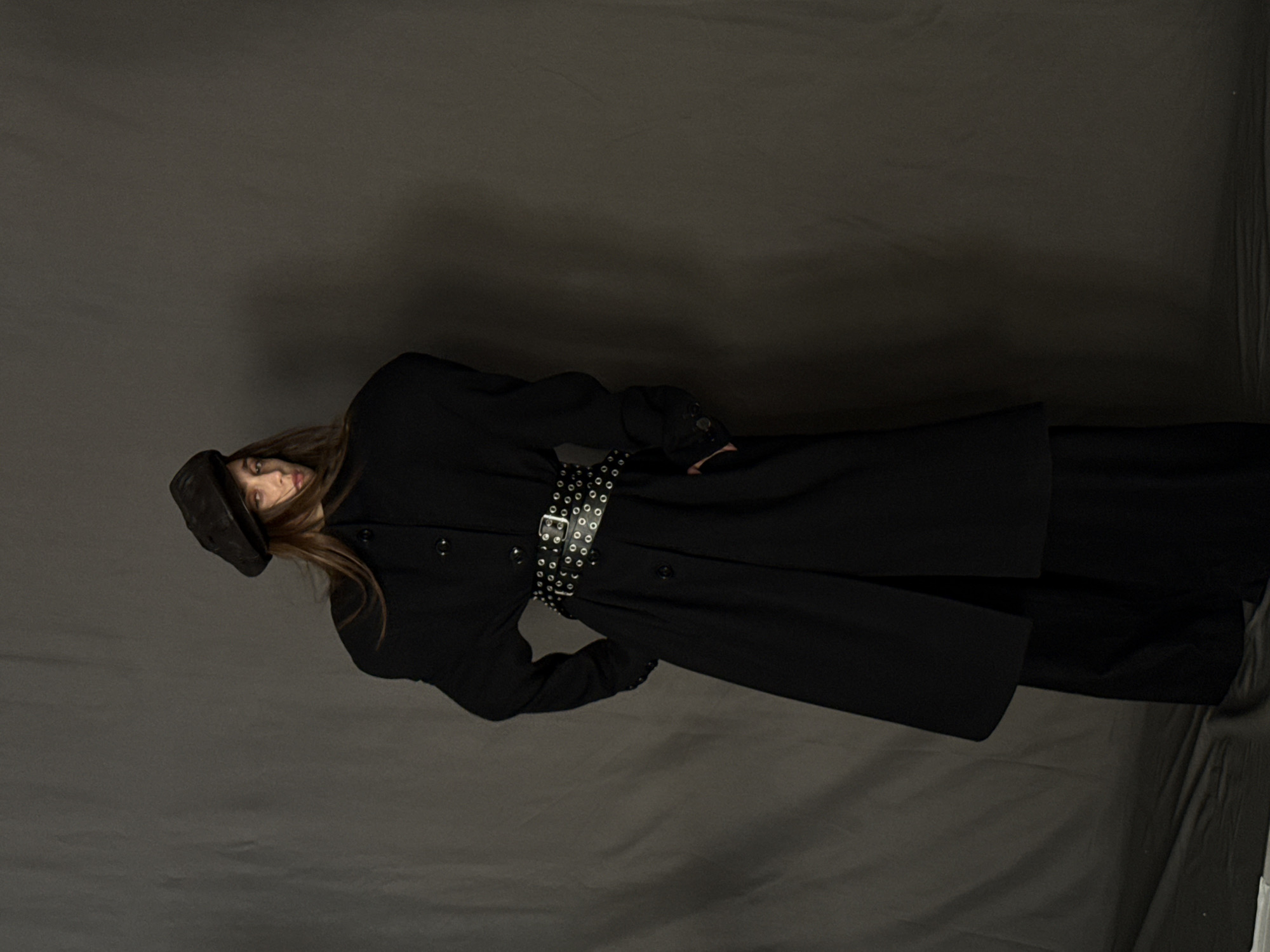 A woman stands against a grey backdrop, wearing a black hat, black coat, and a studded belt. The environment appears to be a studio setting with simple lighting and a neutral background.