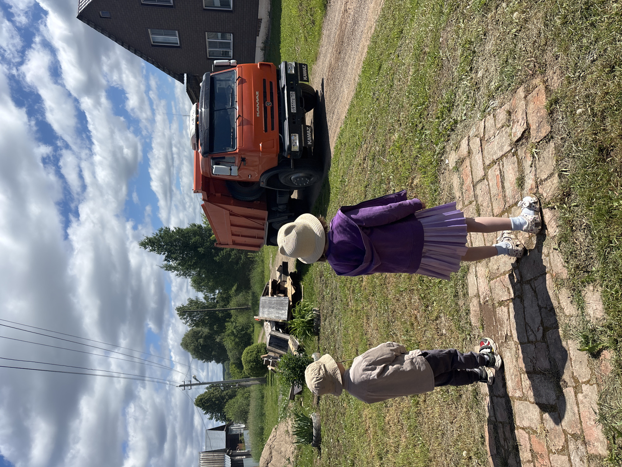 Two children, one wearing a tan hat and beige shirt, the other wearing a white hat and purple jacket with a pleated skirt, stand on a brick pathway facing a large orange dump truck. The truck, a KAMAZ model, is parked on a dirt road. The scene takes place outdoors in a grassy area with trees and houses in the background. Power lines are visible overhead. The overall setting appears to be a rural residential area, possibly in the village of Niva, Russia.