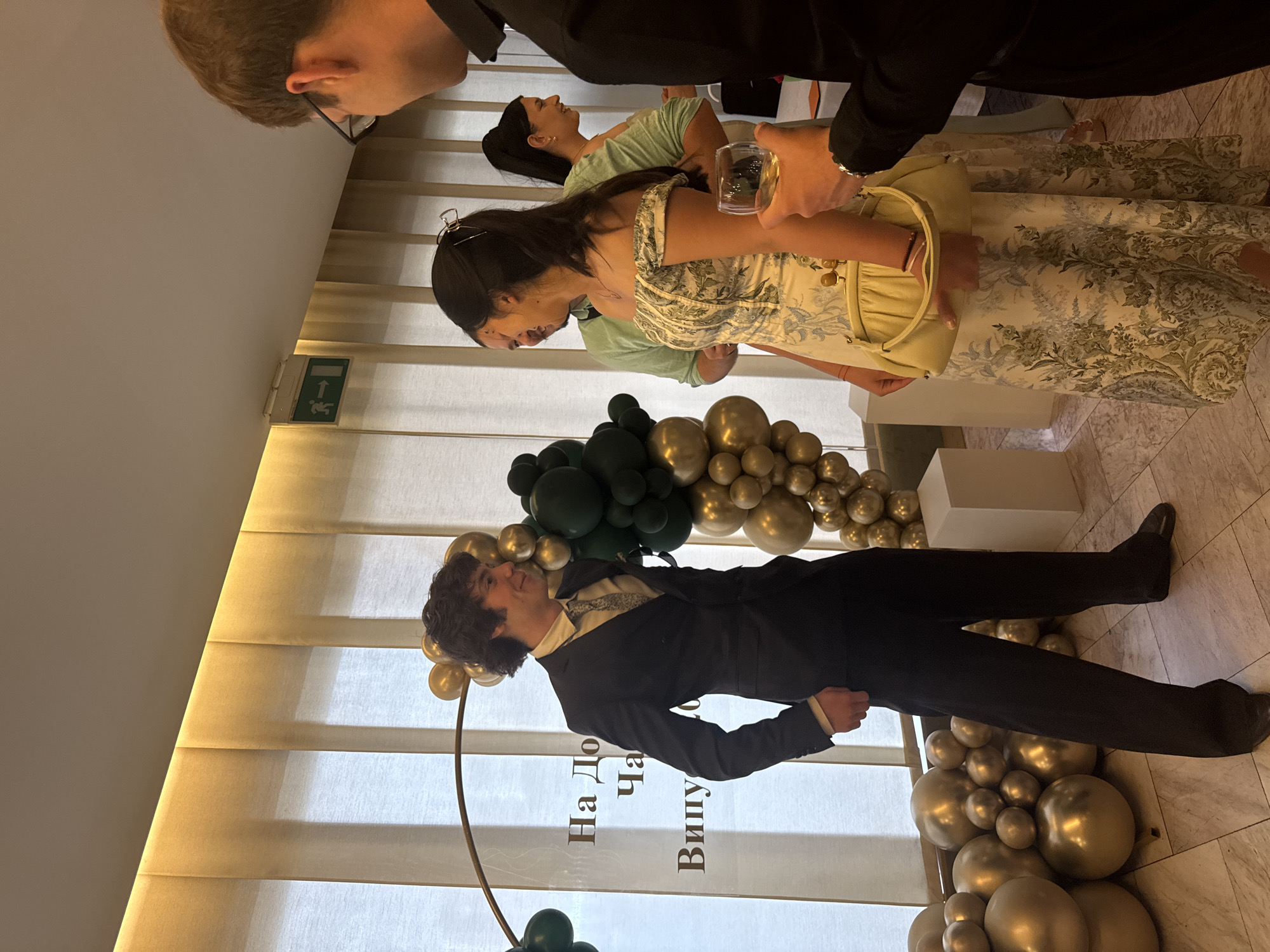 The image captures an indoor celebratory event. A man in a dark suit and tie poses near a balloon arch of green and gold balloons. A clear sign with Cyrillic text is partially visible in front of him. Several other individuals are in the background, including a woman in a floral dress holding a glass, and others engaged in conversation. The event appears to be taking place in a modern, well-lit space with light-colored walls and tiled flooring. Based on the Cyrillic text, this event is likely taking place in Sofia, Bulgaria.