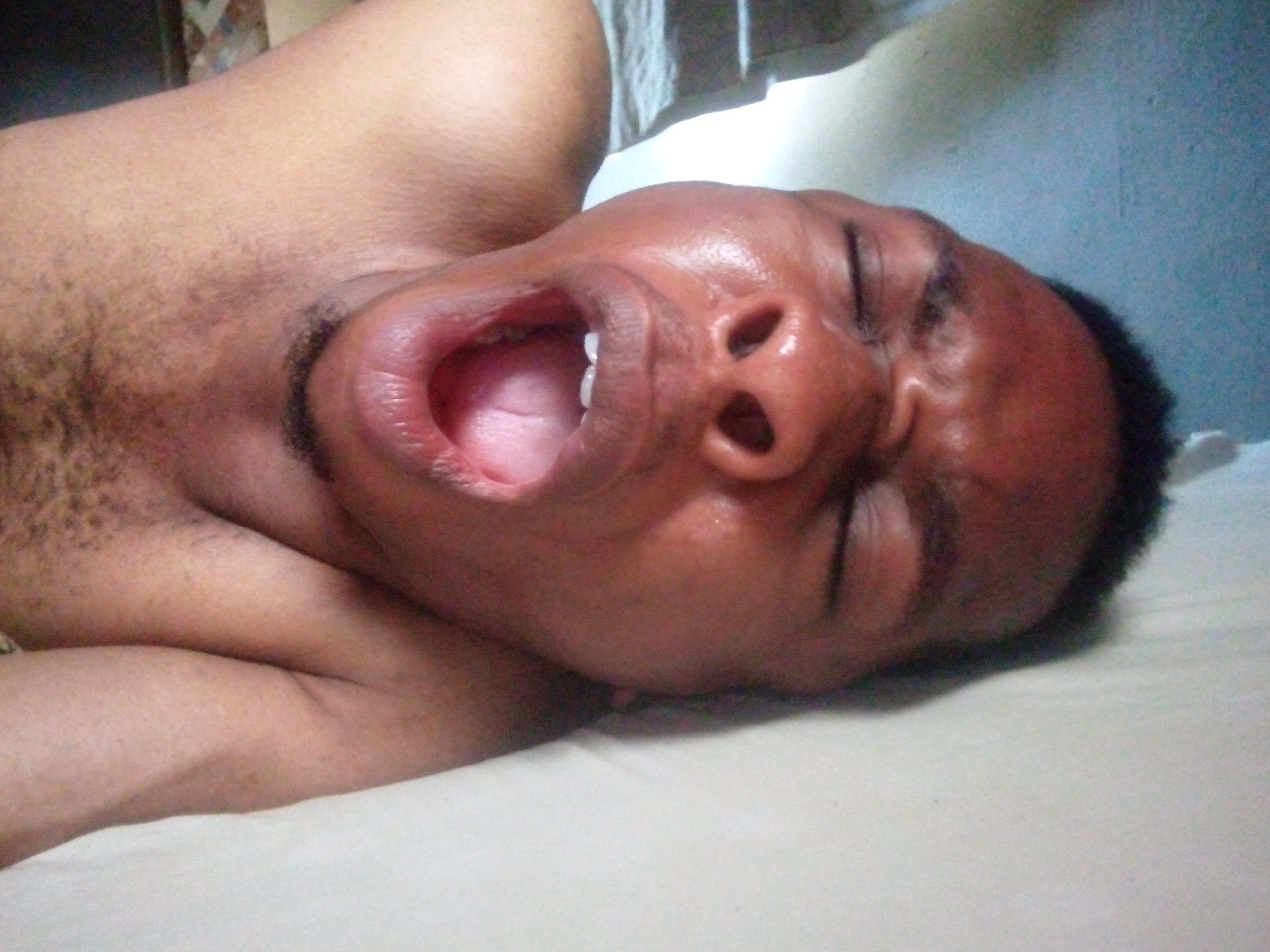 The image depicts a shirtless Black male lying on a white surface. He is lying on his side with his eyes closed and his mouth open wide. His skin appears sweaty. The background is blurry and indistinct, with a hint of light blue on the right and what appears to be a white window covering above. The image lacks location context and does not provide specific details about the city or environment.