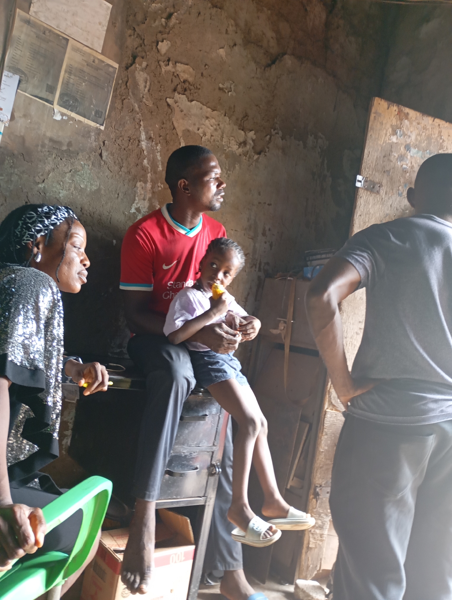 In a dimly lit, rustic interior with rough, textured walls and a wooden door, a family of four is gathered. The scene suggests a humble dwelling, possibly in Shendam, Nigeria. A man in a red Liverpool jersey sits holding a young girl who is eating corn. Next to him, a woman wearing a sparkly silver top and a headscarf looks towards the man. In the foreground, a bright green plastic chair is partially visible. To the right, a man in a grey t-shirt stands with his back to the camera, his arm on his hip. The lighting indicates it is daytime. The overall atmosphere appears relaxed and familial, with the child focused on her snack and the adults engaged in conversation. On the wall behind the man in the red shirt, there are several framed documents, possibly certificates or official papers. The rough texture of the walls and the simple furnishings point to a non-affluent setting.