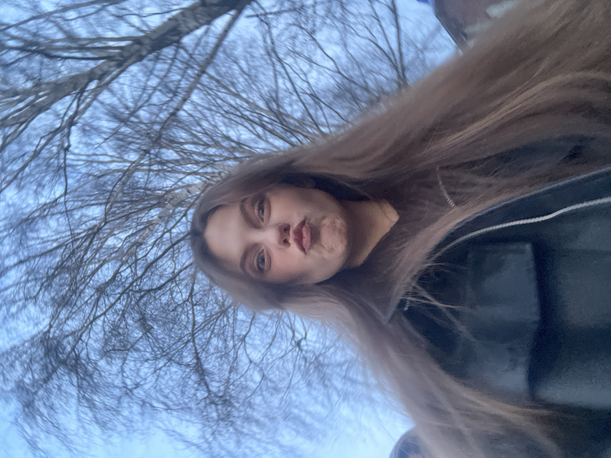 A young woman poses for a low-angle selfie outdoors, with bare trees and a cloudy sky in the background. She has long, light brown hair, light skin, and is wearing a dark jacket with white piping. Her lips are pursed. The location cannot be determined to be Фоки, Russia.