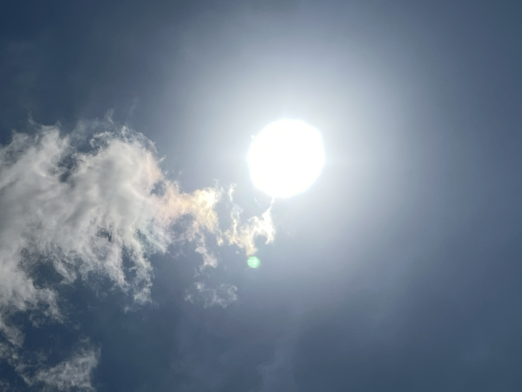 The image shows a bright sun partially obscured by wispy clouds in a clear blue sky. There is a green lens flare visible on the left side of the image, near the clouds. The sun is a large, bright white orb on the right side of the image, with a halo of light emanating from it. The sky is a gradient of blue, darker at the top and lighter towards the bottom. No specific location in Denpasar, Indonesia can be identified from this image.
