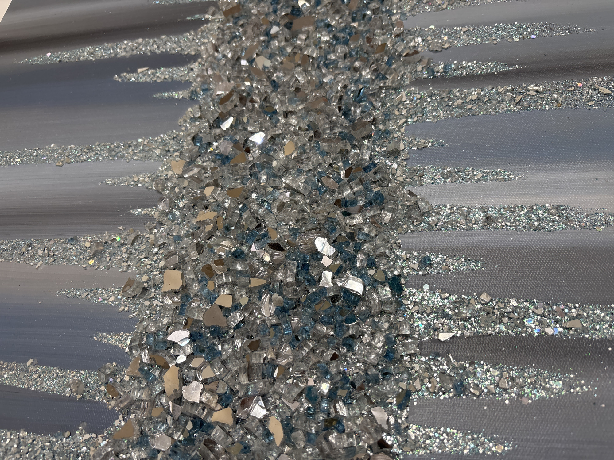The image displays a close-up view of a textured artwork. The artwork features a thick horizontal band of crushed glass and mirror fragments, predominantly in shades of silver, clear, and light blue. This band is applied over a background of vertical, blended strokes of light gray and pale blue paint. Dripping down from the horizontal band are vertical streaks composed of finer glitter and smaller fragments of the crushed glass and mirror, creating a textured, cascading effect. The overall impression is one of sparkle and dimensionality, with the light reflecting off the numerous small surfaces. The image is cropped tightly, focusing solely on the artwork itself, with no discernible environmental context or location information.