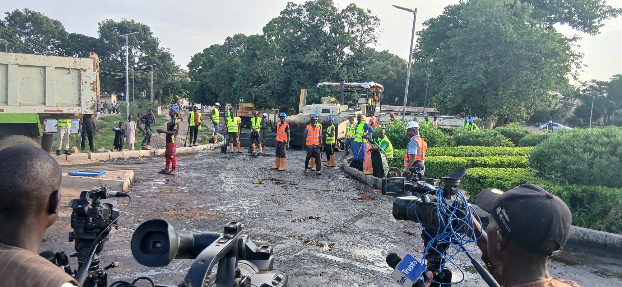 In Jama A, Nigeria, an outdoor scene depicts road construction in progress. A large dump truck, partially visible on the left, is unloading material onto a street that is covered in asphalt and debris. Several construction workers, predominantly male, are actively engaged in the work, many wearing high-visibility vests in yellow and orange, and some with hard hats. They are seen using shovels and operating construction equipment, including a road roller in the background. A few bystanders are also present, observing the activity. The environment is characterized by trees and lush green bushes lining the road, suggesting a well-maintained urban or semi-urban setting. The sky is overcast, indicating diffused natural light. In the foreground, two professional video cameras are set up, suggesting that the event is being documented.