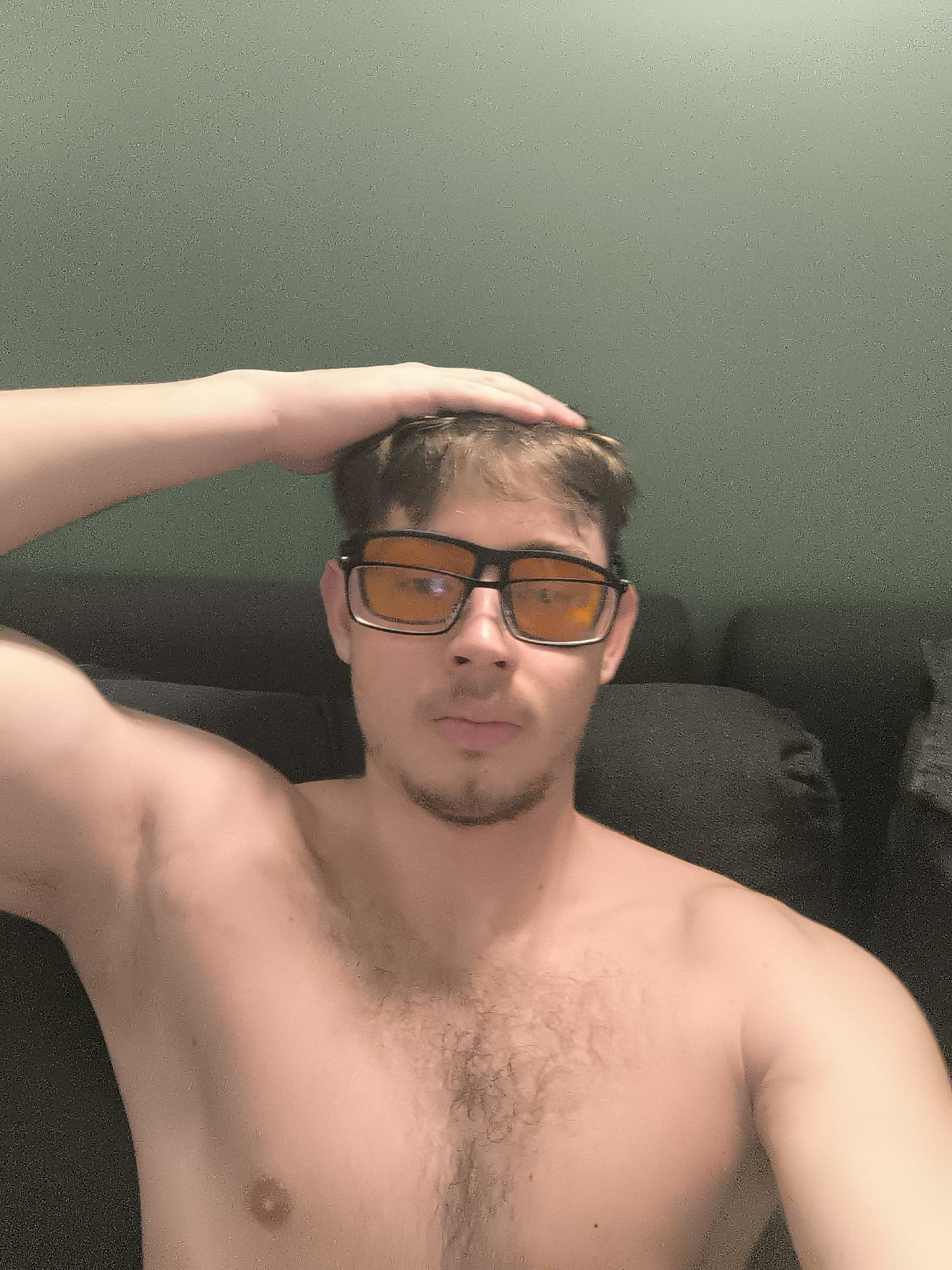 A shirtless man with brown hair and a light beard is wearing orange-tinted glasses. His right hand is resting on his head. He is sitting on a dark gray couch in front of a plain, light green wall. There is no indication of Sofia, Bulgaria.
