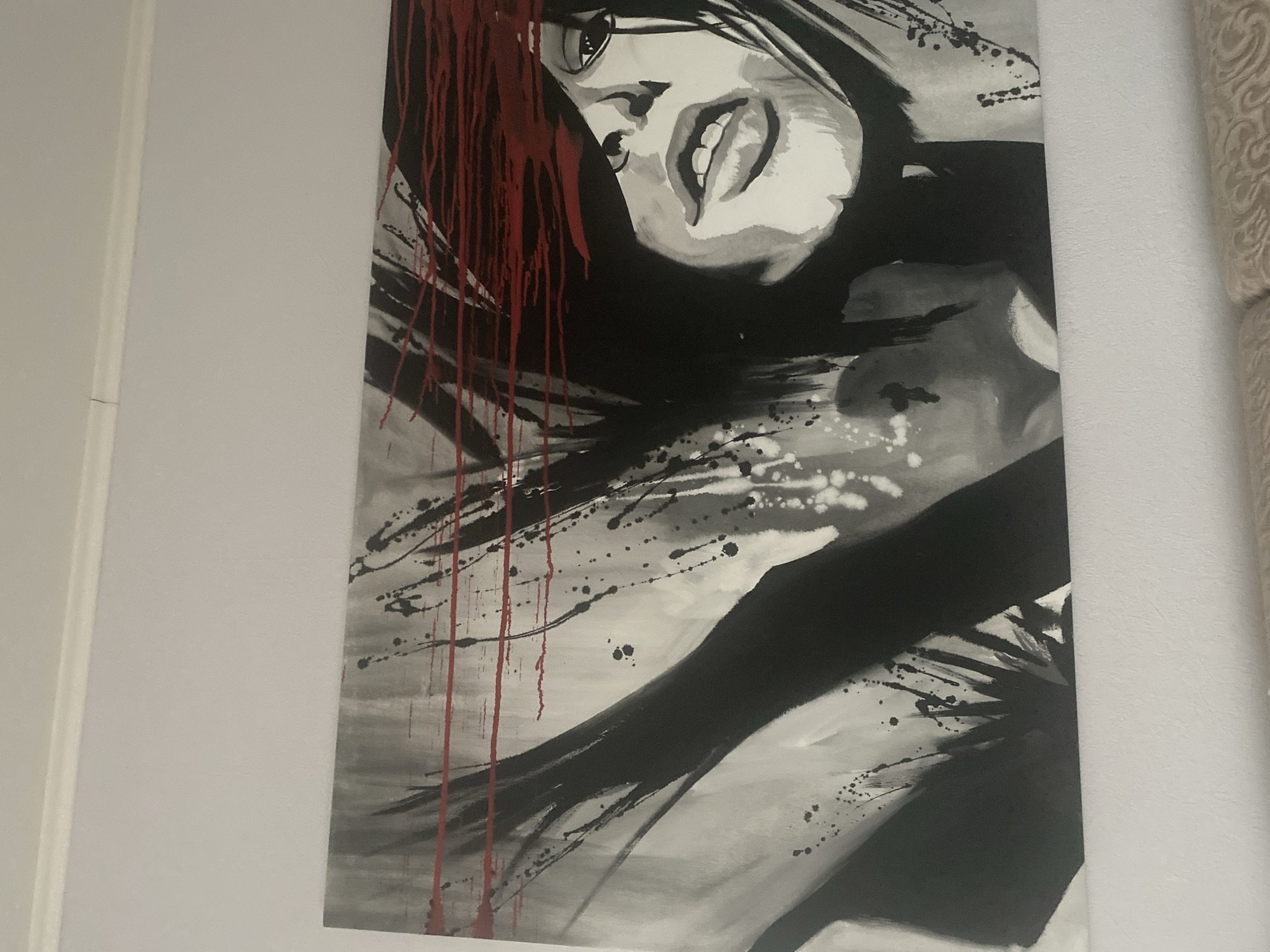 The image displays an interior space featuring a large horizontal canvas art piece mounted on a uniform light-colored wall. The scene does not contain any identifiable characteristics specific to Krupki, Belarus.
The artwork depicts a stylized, partially obscured female face rendered in black, white, and various shades of gray, highlighted by prominent red elements. The lower portion of the face, encompassing the nose, an open mouth revealing teeth, and the chin, is visible, conveying an intense expression. The upper facial area, including the eyes, is completely obscured by multiple horizontal streaks of vibrant red paint that extend across the canvas. Dynamic, dark, abstract brushstrokes, suggestive of flowing hair or energetic movement, frame the visible facial features. The background layers various gray tones with scattered black paint splatters and drips, contributing to a sense of motion.
A white crown molding runs along the upper perimeter of the wall. A section of light-toned, textured furniture, displaying a subtle patterned design, is visible at the bottom of the frame. No individuals are physically present within the depicted room; the human form is solely represented within the artwork.