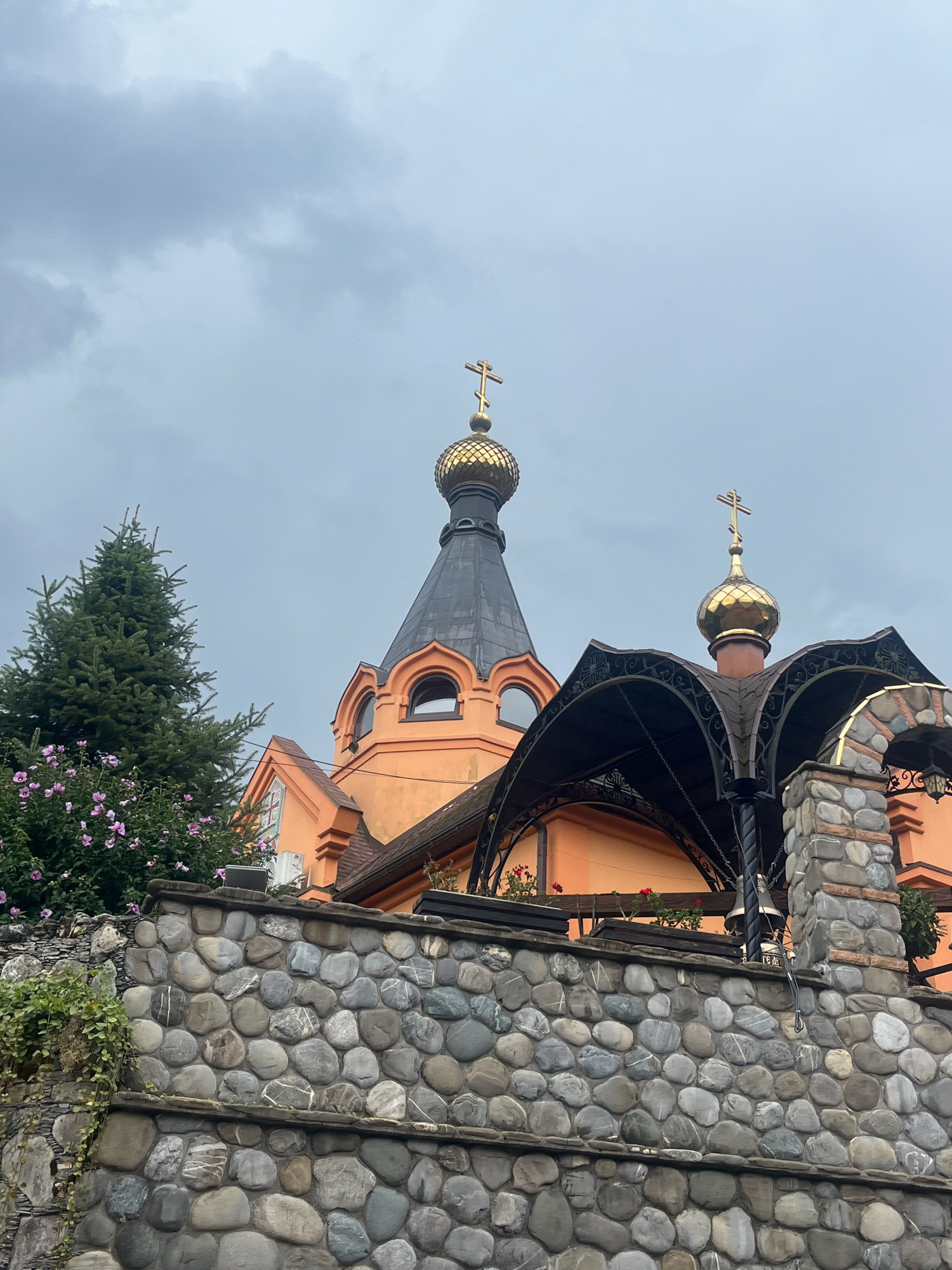 An orange-colored building with gold-domed roofs and crosses is partially visible behind a grey stone wall in Solokh-Aul, Russia. A large green coniferous tree stands to the left of the building, with pink flowers blooming on a bush in front of it. The sky is overcast. The stone wall in the foreground is constructed of large, smooth, rounded stones. A decorative metal canopy extends over a portion of the building.