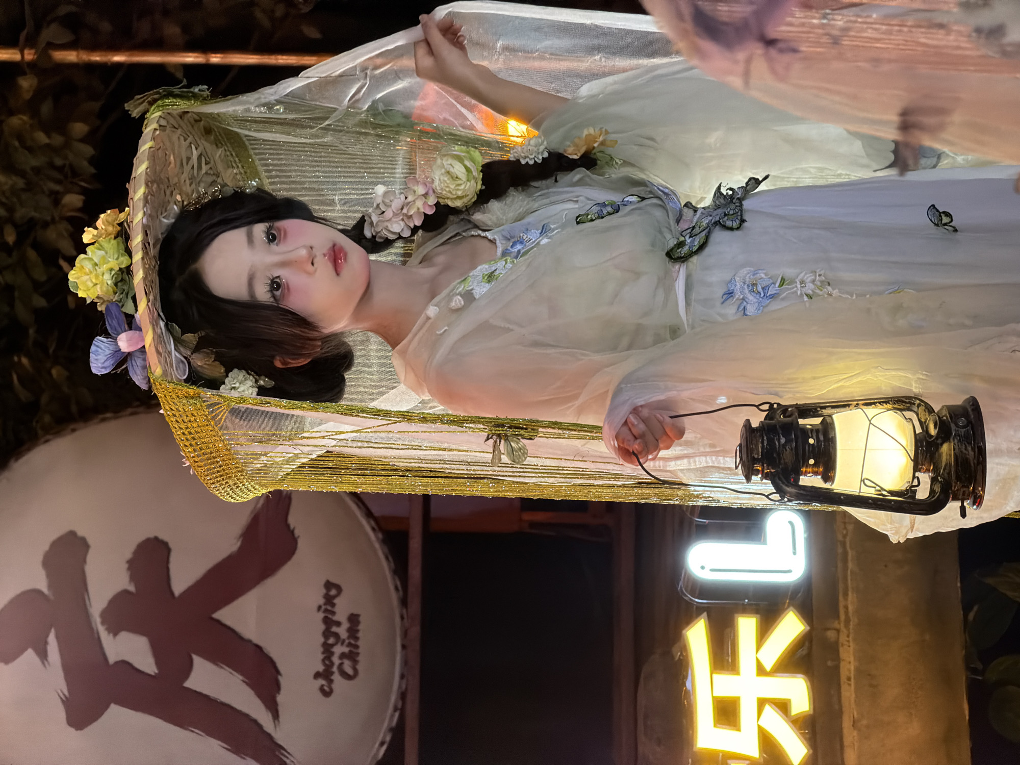 A young East Asian woman in traditional Chinese attire stands in front of a large drum with Chinese characters painted on it. She wears a light-colored, flowing garment adorned with floral embroidery and a wide-brimmed hat decorated with flowers and netting. She holds an old-fashioned lantern in her right hand. Behind her, neon signs that read "乐 L" are visible. The scene appears to be outdoors at night, possibly in a commercial or cultural area. The drum has the text "chongqing China" below the large characters.