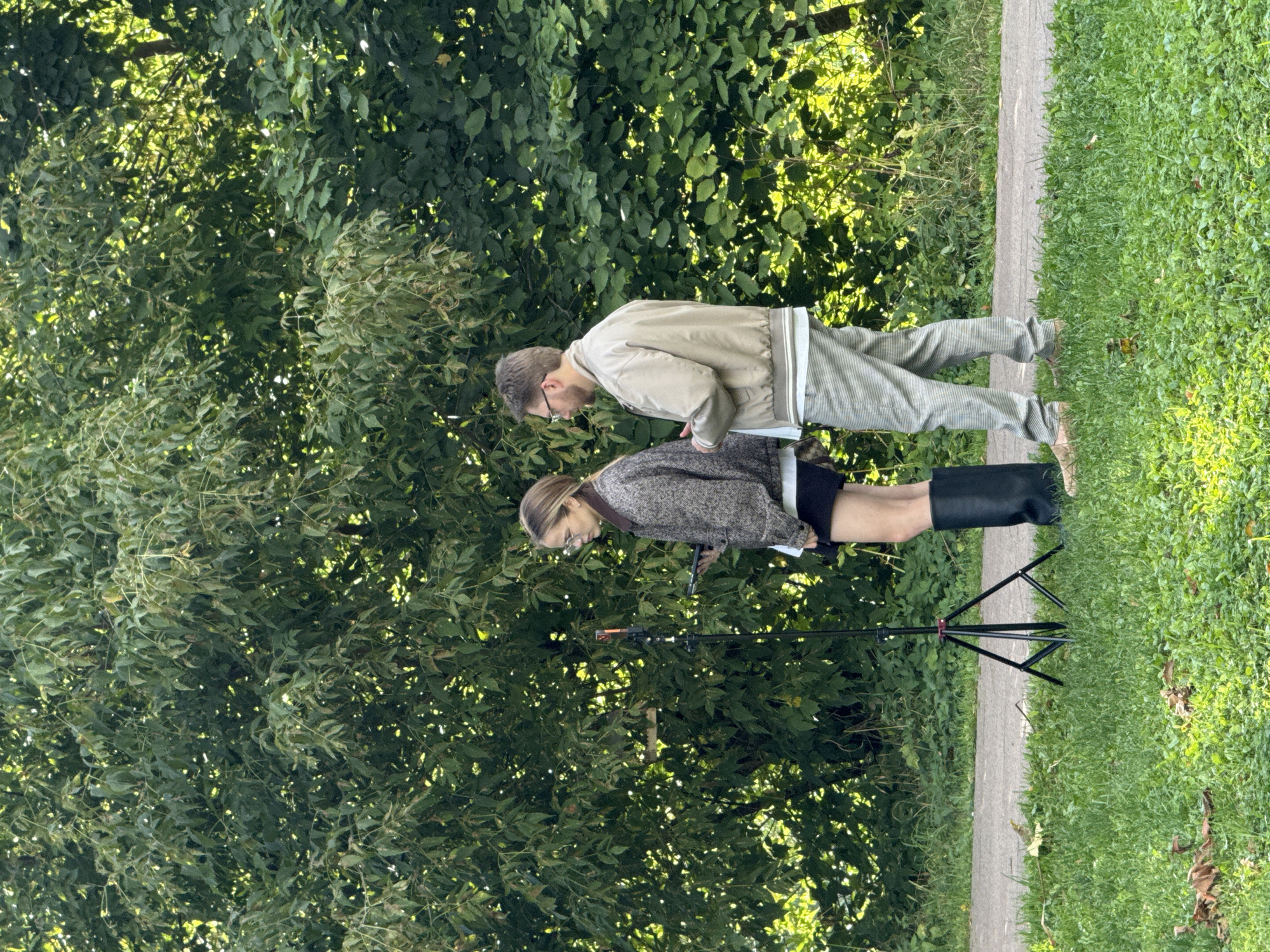 Two individuals, a woman and a man, stand outdoors near a treeline. The woman, on the left, wears a tweed jacket, a dark skirt, and tall black boots. She holds a tablet in her hands. To her right, a tripod supports an orange device. The man, on the right, wears a light-colored bomber jacket, a striped shirt, and light-colored pants. He also holds a tablet. They appear to be engaged in an activity that involves the devices and possibly the tripod-mounted object, suggesting a filming or recording session. The background consists of lush green foliage from trees and shrubs, with a paved path visible behind the tripod and grass in the foreground. This scene is located in Dolgoprudny, Russia.