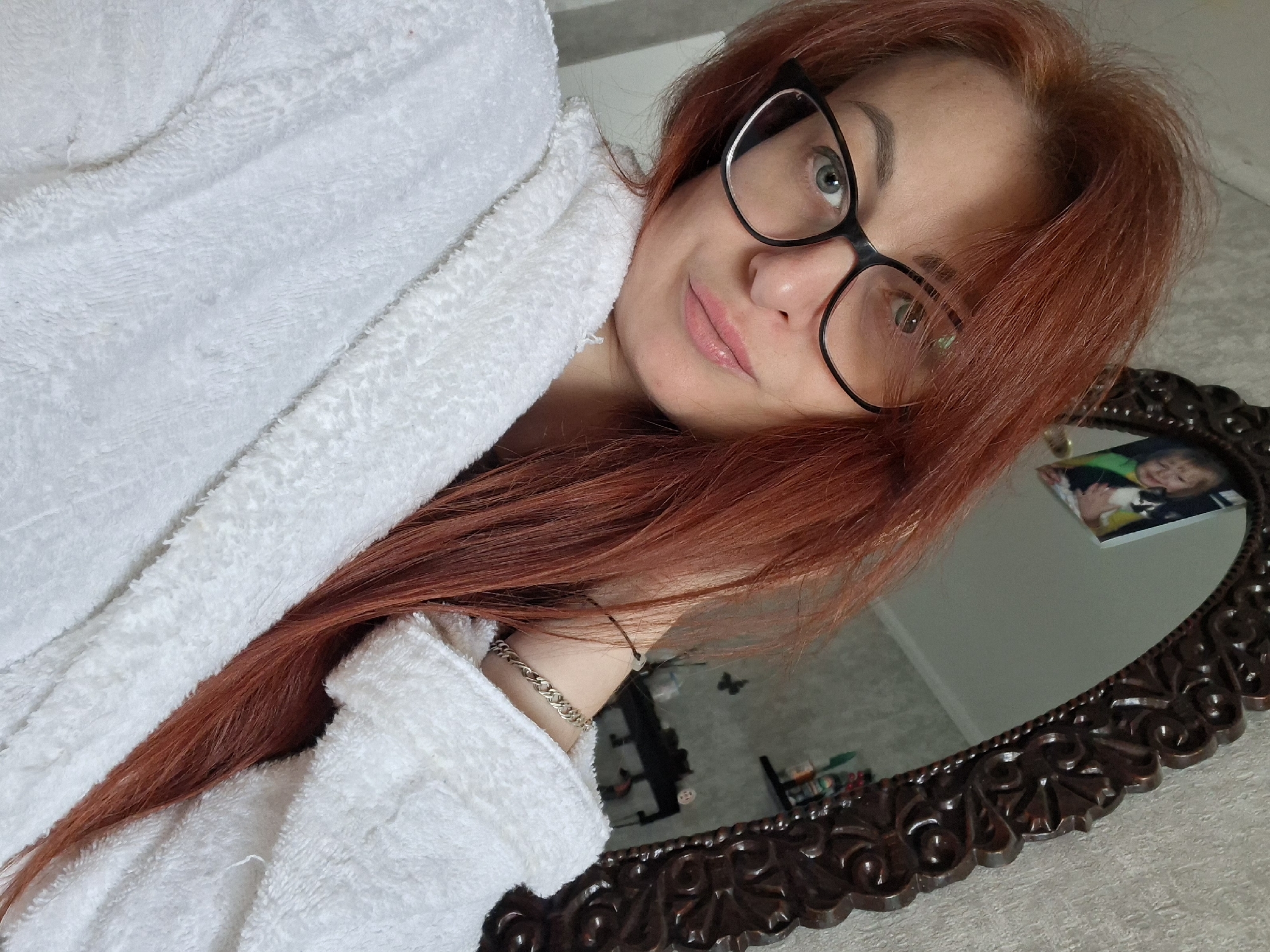 A woman with long, reddish-brown hair and glasses is taking a selfie. She is wearing a white bathrobe and has her hand on her neck. In the background, a decorative mirror reflects a picture of a child holding a cat. The scene appears to be indoors, possibly in a bedroom or bathroom, with textured wallpaper visible.