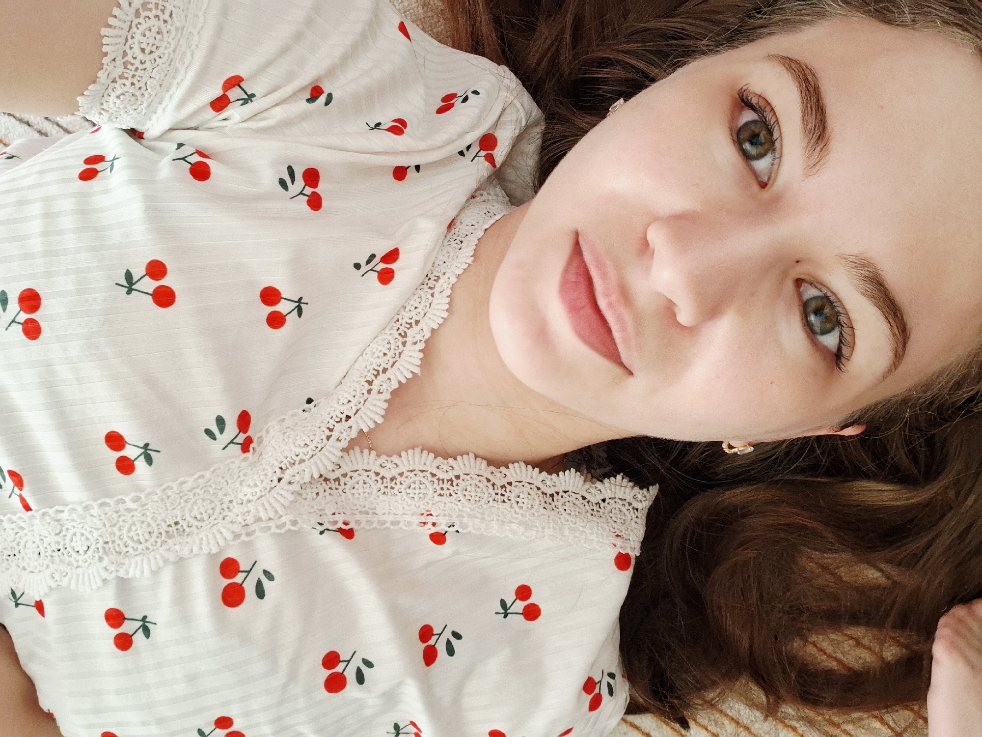 A close-up, slightly angled, indoor shot features a young woman lying down, presumably indoors. She is wearing a white top adorned with a cherry pattern and lace trim. Her hair is brown and wavy, and she has light skin, brown eyebrows, and hazel eyes. She is smiling slightly. A small earring is visible in her right ear. Part of a hand is visible in the lower right corner. The background appears to be a bed or similar soft surface. There's no explicit evidence to confirm or deny the location in Chaikovsky, Russia.