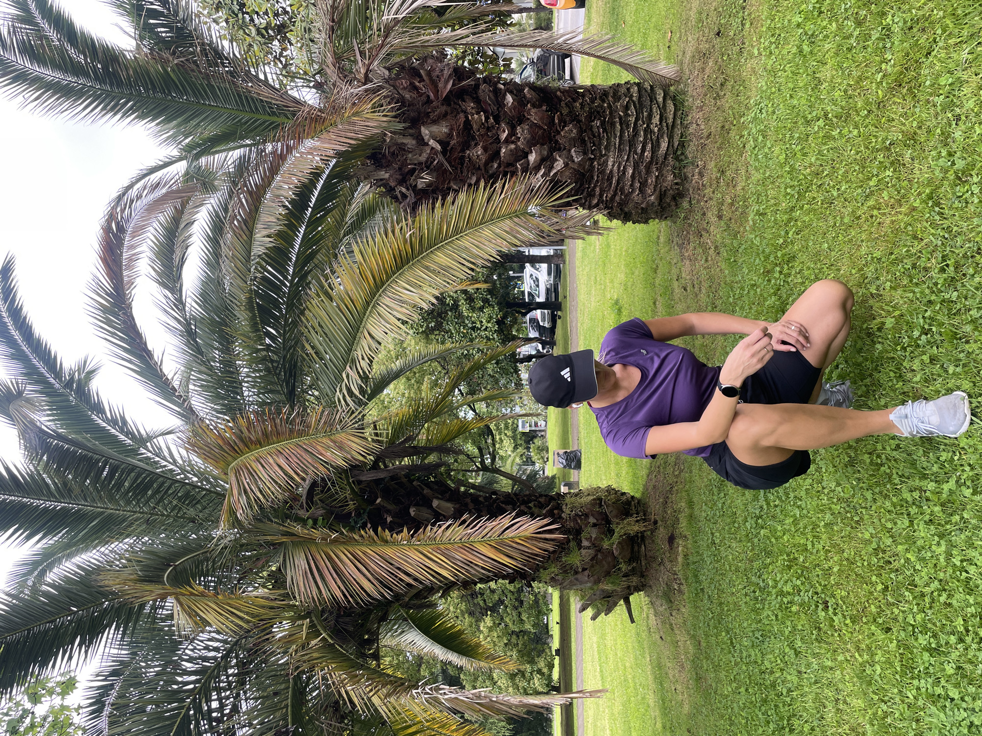 A woman in a purple shirt, black shorts, and a black Adidas cap is crouching on grass next to a palm tree. She is wearing grey running shoes and a smartwatch on her left wrist. The palm tree has brown trunk and green and yellow fronds. Behind the palm tree, there are other trees and a few cars parked in the distance. The ground is covered in green grass. The picture was taken in Sochi, Russia.