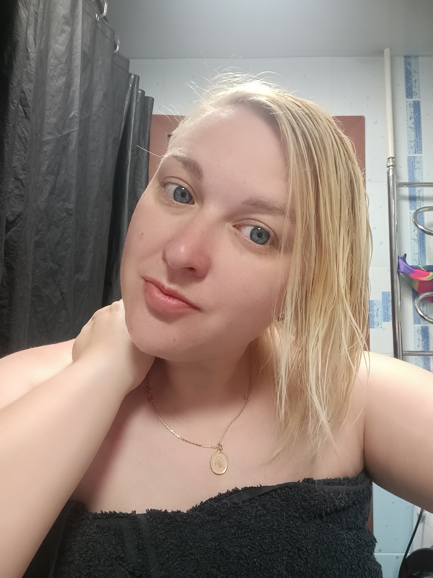 A woman with short, wet, light blonde hair is in a bathroom. She is wearing a black towel wrapped around her torso and a gold necklace with a round pendant. Her skin is fair, and she has light blue eyes. She has her right hand on her neck and is looking at the camera with a slight smile. Behind her is a gray shower curtain on the left and light blue-and-white tiled walls with a towel rack on the right. A door is visible above the towel rack. The location appears to be a residential bathroom.