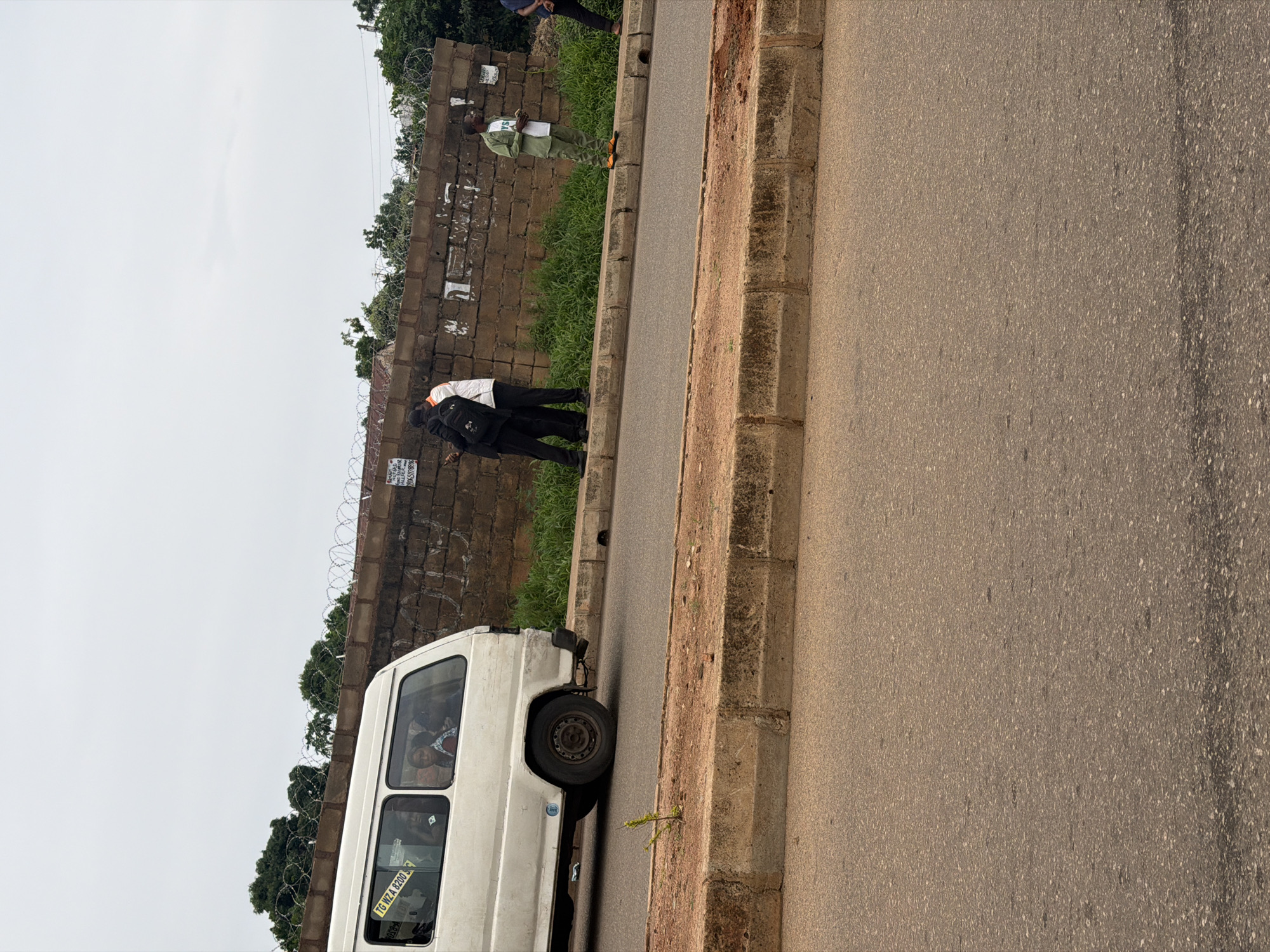 A white passenger van, with the license plate "TG WZA 8200 S", is parked on the left side of a two-lane paved road. Several people are visible inside the van, looking out the windows. On the right side of the road, a man in a green uniform and white t-shirt stands near a brick wall topped with barbed wire. Behind him, two men, one in a white shirt and dark pants, and the other in dark clothing with a backpack, are standing close together. The environment appears to be a roadside in a city, possibly Bukuru, Nigeria, with a concrete curb and vegetation bordering the road and wall. The sky is overcast.