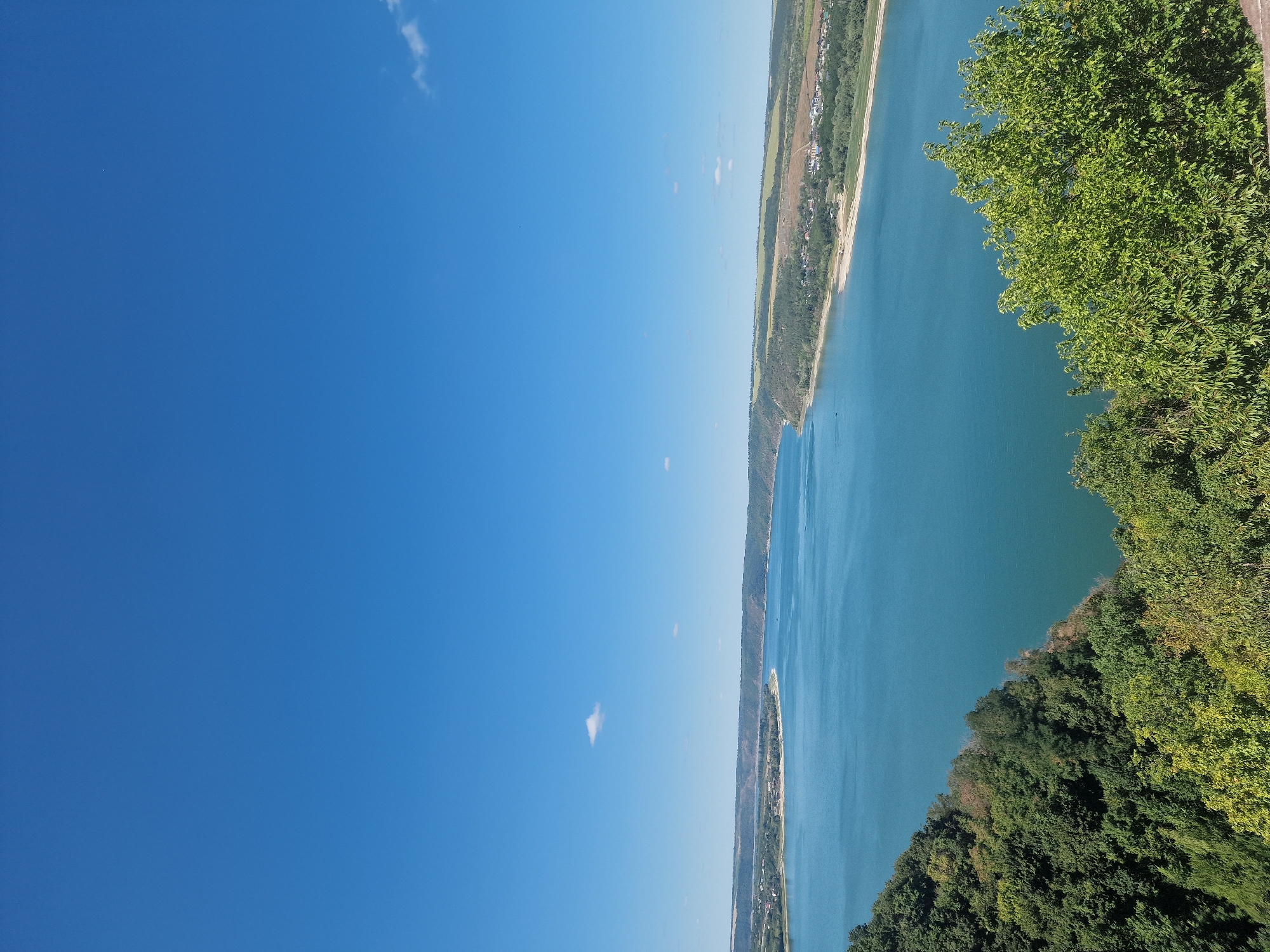 This is an aerial, wide-angle shot capturing a vast, calm body of water, likely a river or lake, with a bright blue sky overhead. The water occupies the majority of the frame, appearing a deep turquoise, with its surface relatively smooth. Along the far bank of the water, a low, rolling landscape unfolds, featuring a mix of green vegetation, agricultural fields, and scattered buildings forming a small settlement or town. The shoreline is visible, with a sandy or light-colored beach in places.
In the foreground, the image is dominated by lush green foliage, dense trees, and undergrowth, suggesting a hillside or elevated viewpoint overlooking the water. The vegetation in the foreground is a vibrant green, indicating a healthy and flourishing natural environment. The sky is a clear, bright blue with a few wispy white clouds scattered sparsely, contributing to the overall serene and bright atmosphere. There are no people or visible animals in the scene. The perspective is from a high vantage point looking down and across the water. The location is not identifiable as Saint-Josse-ten-Noode - Sint-Joost-ten-Node, Belgium; the landscape depicted is not characteristic of that urban Belgian municipality.