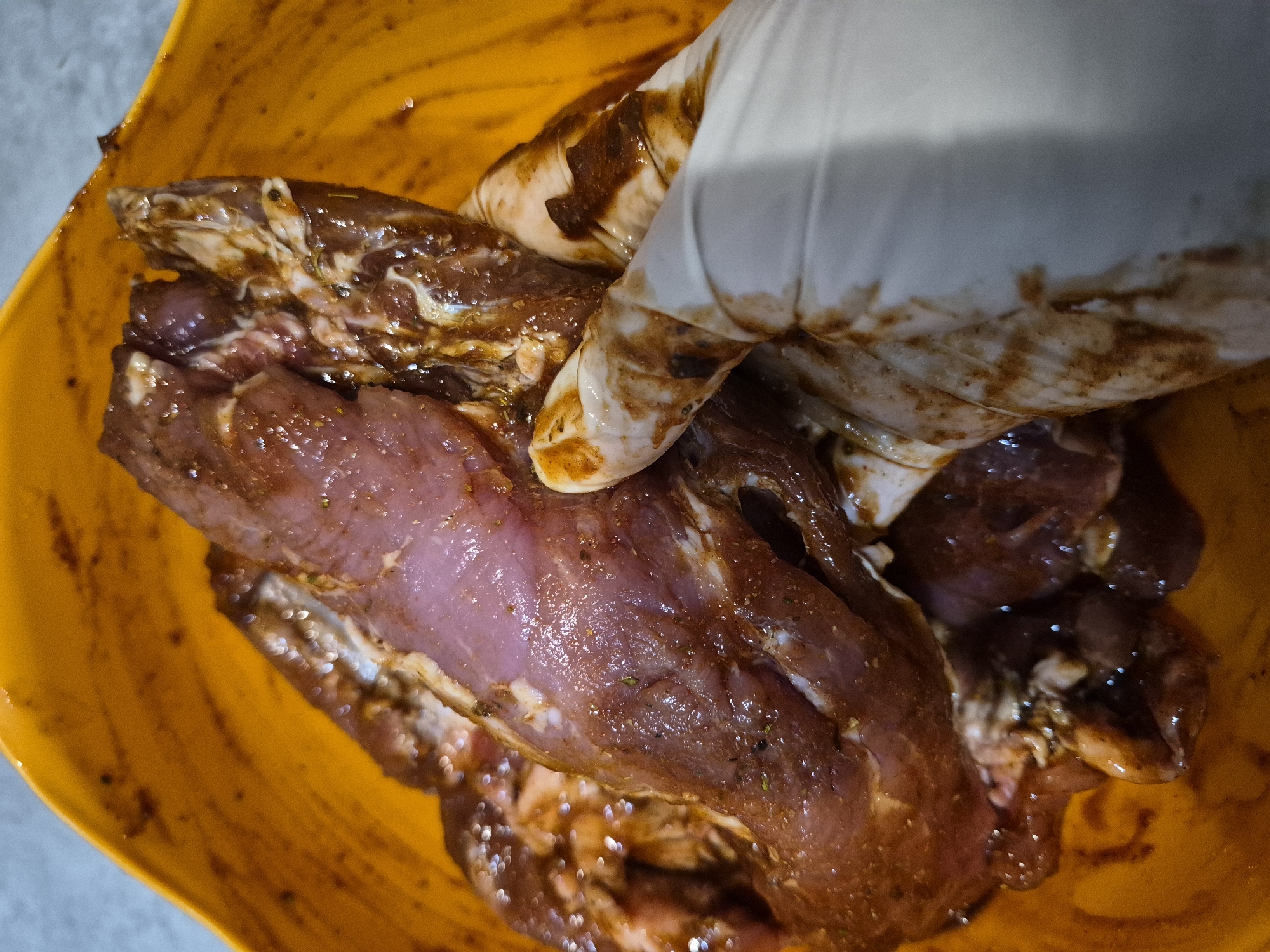 The image shows a close-up view of raw meat being marinated in a bright orange bowl. A hand wearing a white glove is visible, actively massaging the marinade into the meat. The meat appears to be a cut of pork tenderloin, with visible streaks of fat and a pinkish-purple hue. The marinade is dark brown and has a glossy sheen, coating the meat and the inside of the bowl. The texture of the meat is moist and appears to have herbs and spices mixed into the marinade. The background is out of focus, showing a light-colored, textured surface, possibly a countertop. The lighting suggests an indoor setting, likely a kitchen, and the overall impression is one of food preparation. No text is visible in the image.