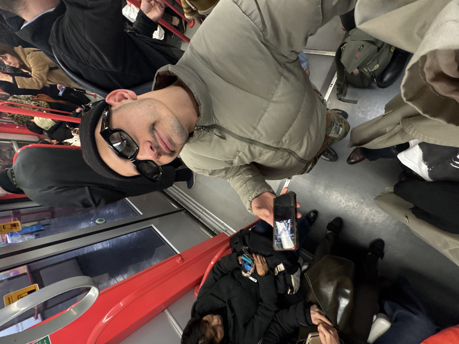 The image captures the interior of a crowded Milan Metro train in Milan, Italy. A male individual is the primary subject in the foreground, looking directly at the camera with a slight smile. He wears a black beanie, black rectangular sunglasses, and a light olive green puffer jacket, and has a visible mustache. He holds a smartphone horizontally in his left hand, its screen reflecting light. To his left, a seated woman with dark hair, wearing a dark coat, holds a phone and other items in her lap. Multiple other passengers are visible throughout the train car; some are standing and gripping red vertical handrails, while others are partially seated. One person in the mid-ground stands with their back to the camera, wearing a long dark coat. The train interior features bright red handrails and supports, silver-framed glass doors, and a gray floor. A green backpack rests on the floor near the main subject's feet. The scene depicts a typical urban public transportation environment with commuters.